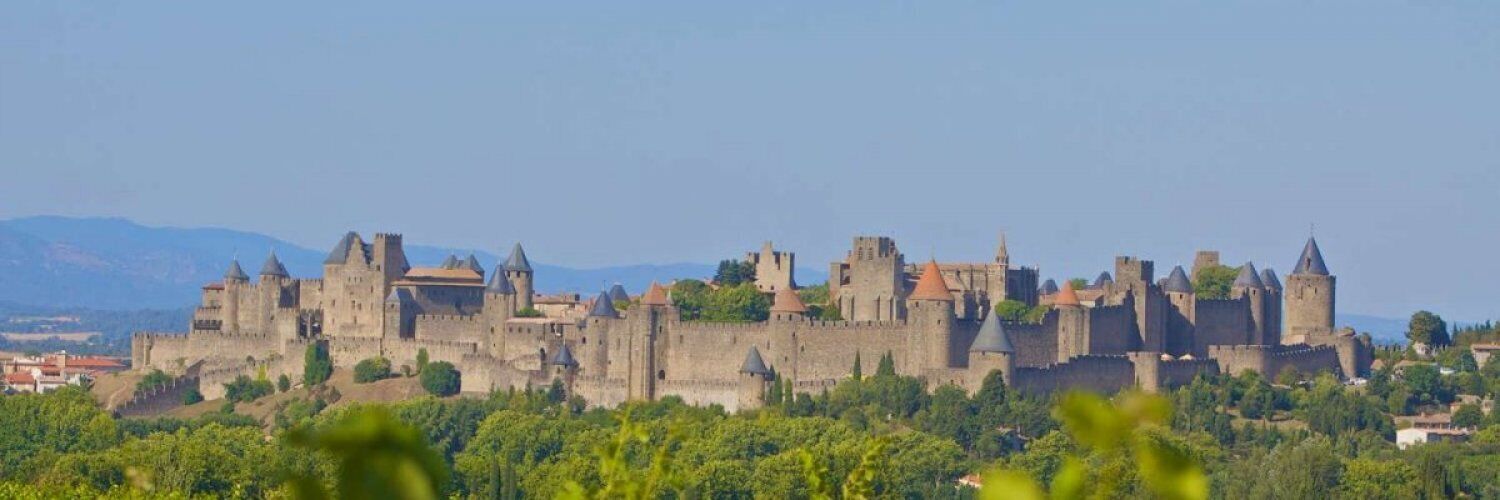 A medieval castle with multiple towers and walls, surrounded by greenery and mountains in the background.