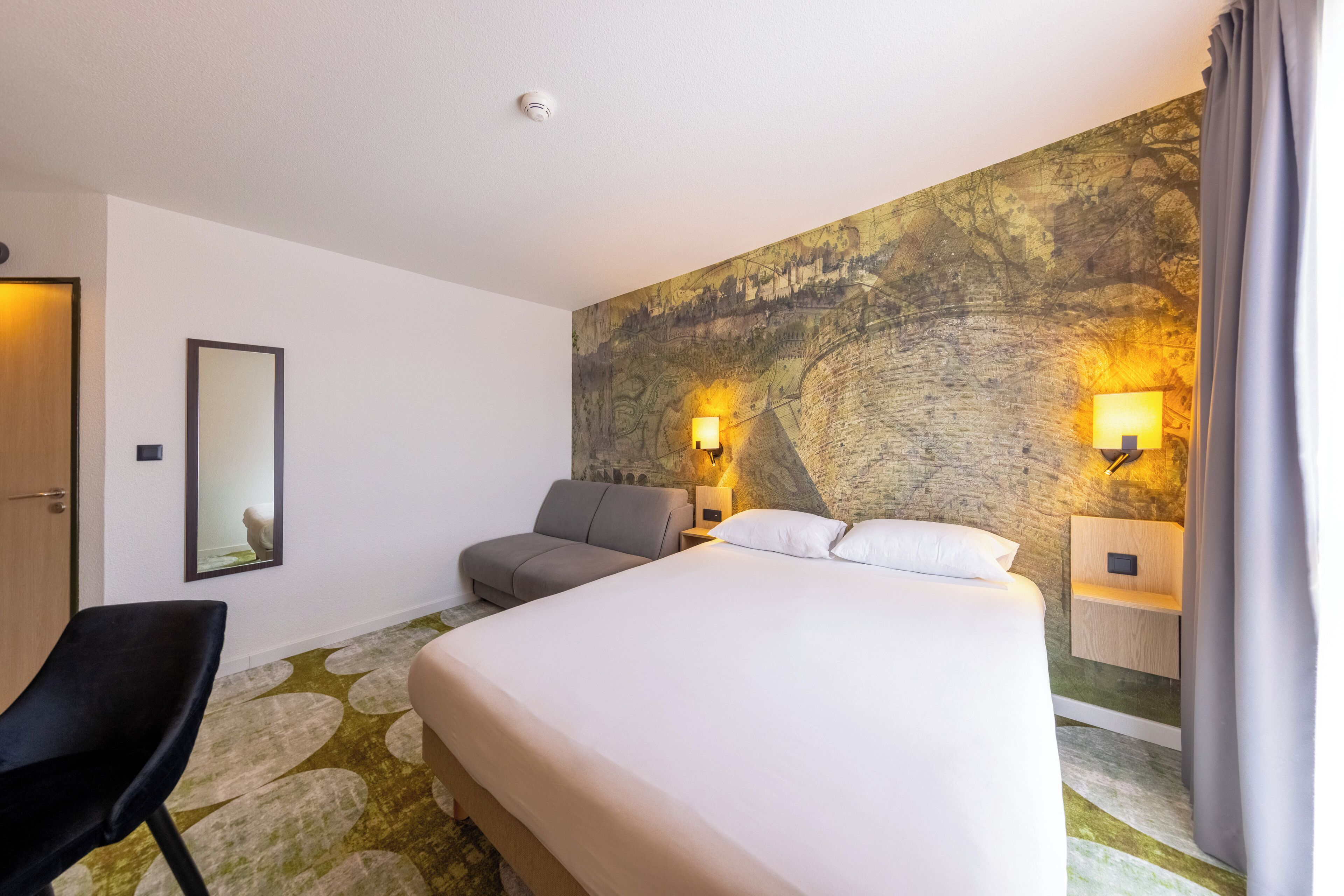 A hotel room with a large bed, a sofa, a chair, and a wall-mounted TV.