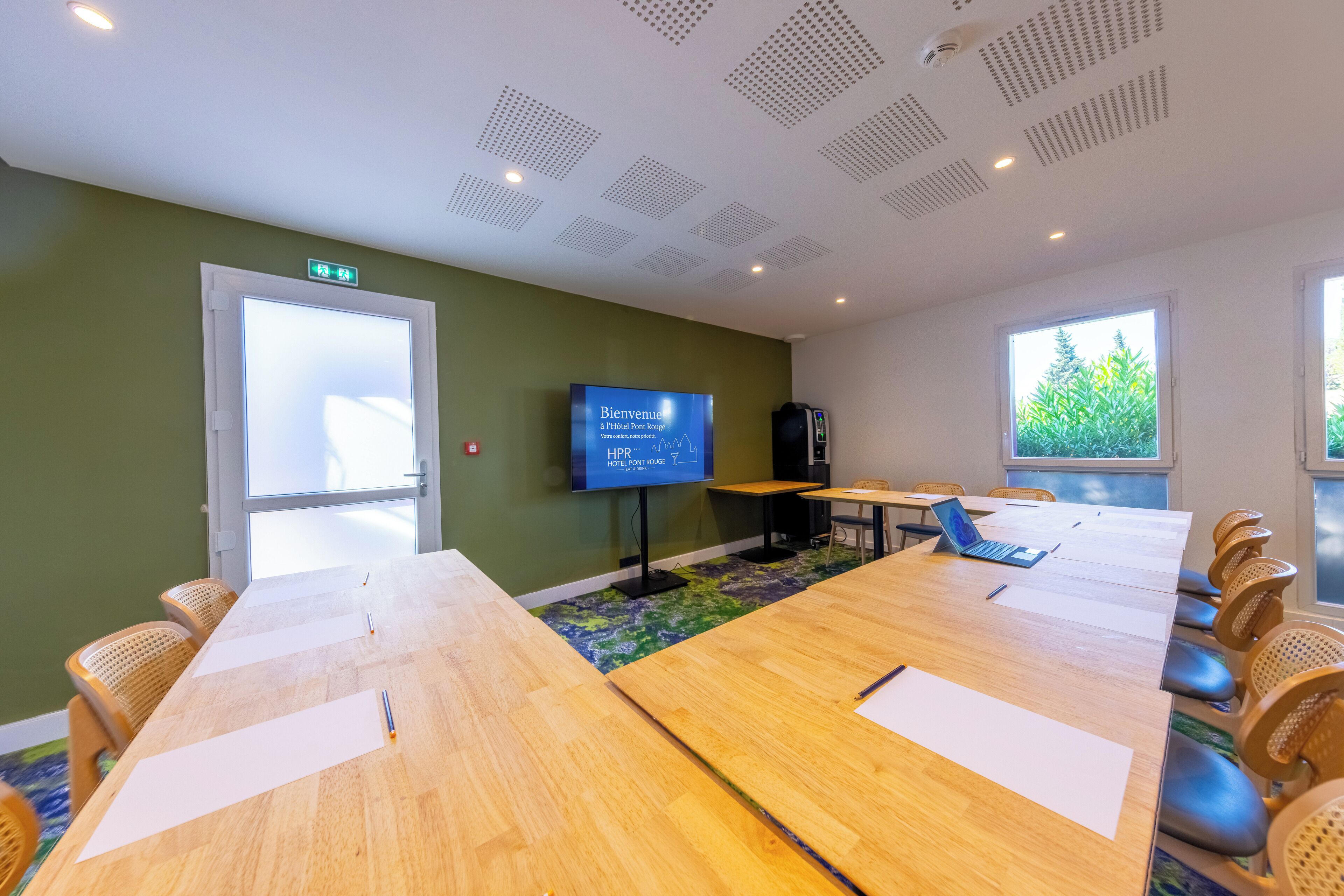 A conference room with a long table, chairs, a television displaying 'Bienvenue & Hotel Point Rouge', and a view of greenery outside.
