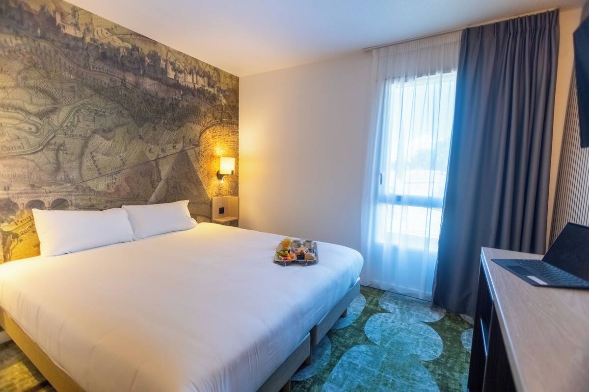 Une chambre d’hôtel avec un grand lit, une carte géographique peinte sur le mur, un bureau avec un ordinateur portable et une fenêtre avec des rideaux.
