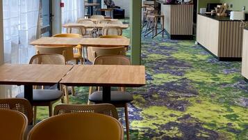 Een moderne eetruimte met houten tafels en stoelen, een receptiebalie en een opvallend groen en paars tapijt.