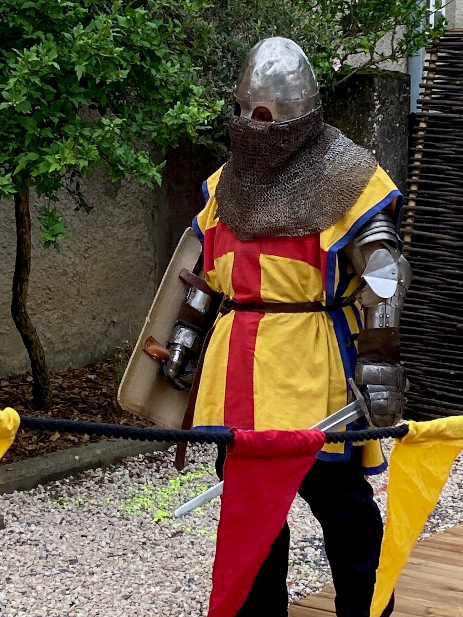A person in medieval armor with a shield and sword, standing outdoors.
