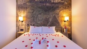A neatly made bed with white linens and red rose petals, two bedside lamps, and a wall mural of a cityscape. - Hôtel Pont Rouge, Carcassonne (Carcassonne)