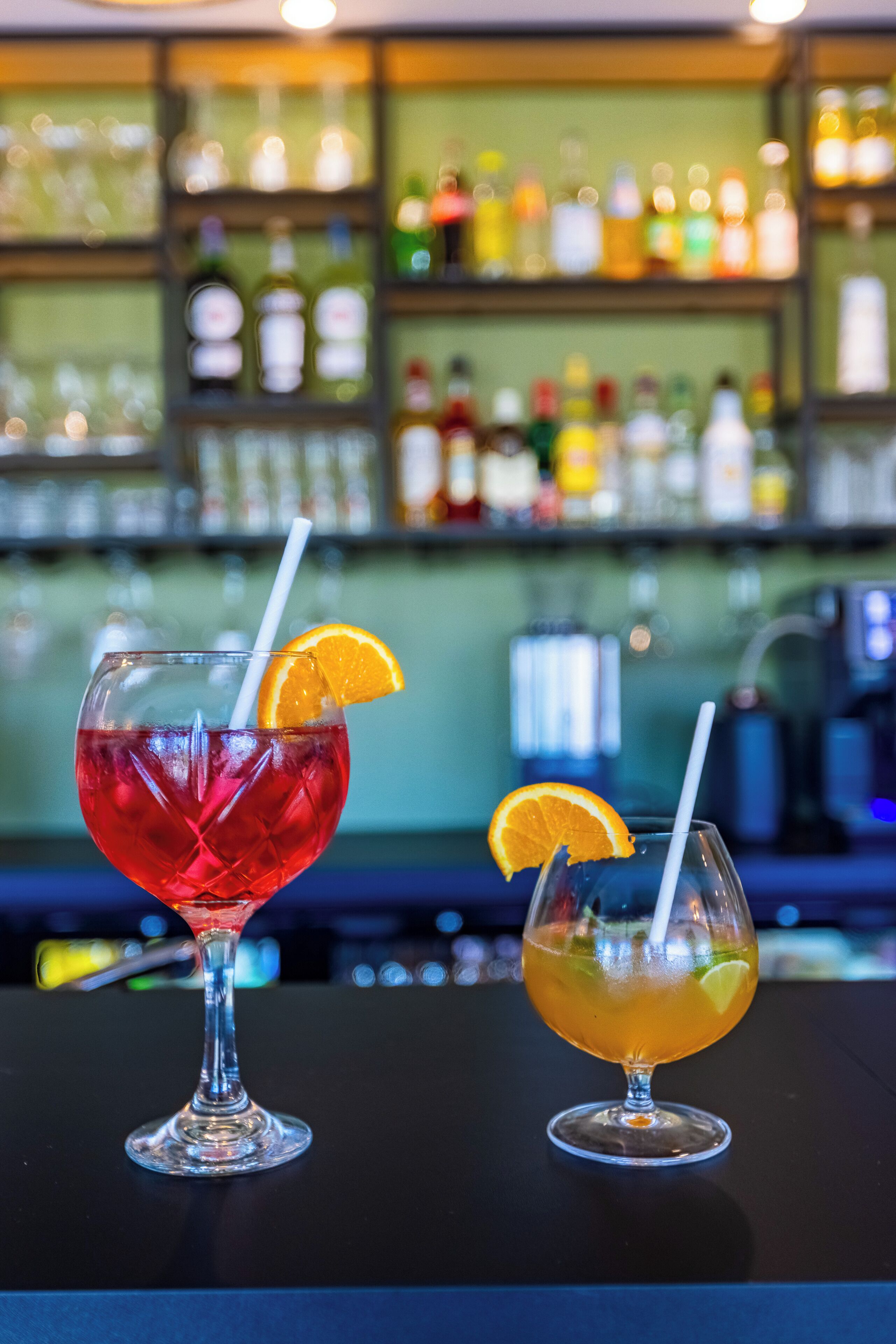 Deux cocktails sur le comptoir d’un bar, avec en arrière-plan, flou, des étagères remplies de bouteilles.
