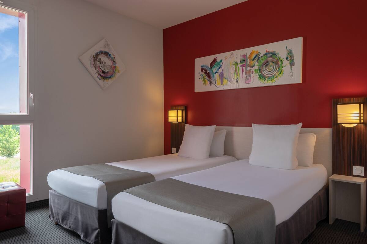 Une chambre d’hôtel avec deux lits, un mur d’accent rouge, des œuvres d’art et une fenêtre donnant sur un paysage agréable.