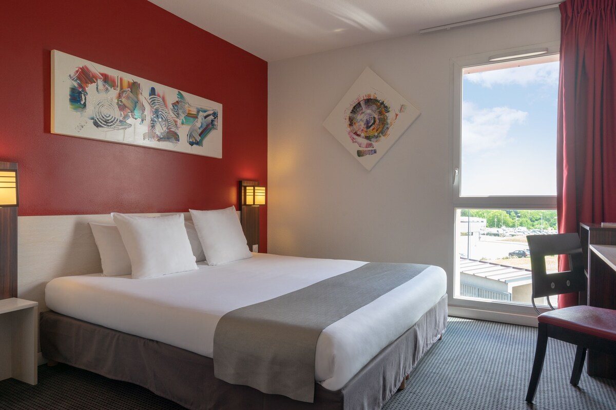 A hotel room with a bed, a desk, a chair, two paintings, and a window with a view.