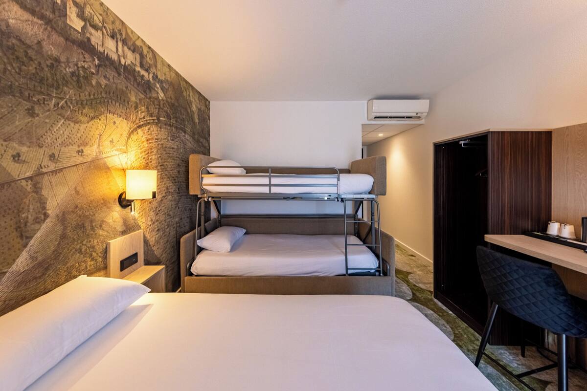 Une chambre d’hôtel avec un lit superposé, un bureau avec une chaise, un climatiseur fixé au mur et une table de chevet avec une lampe.
