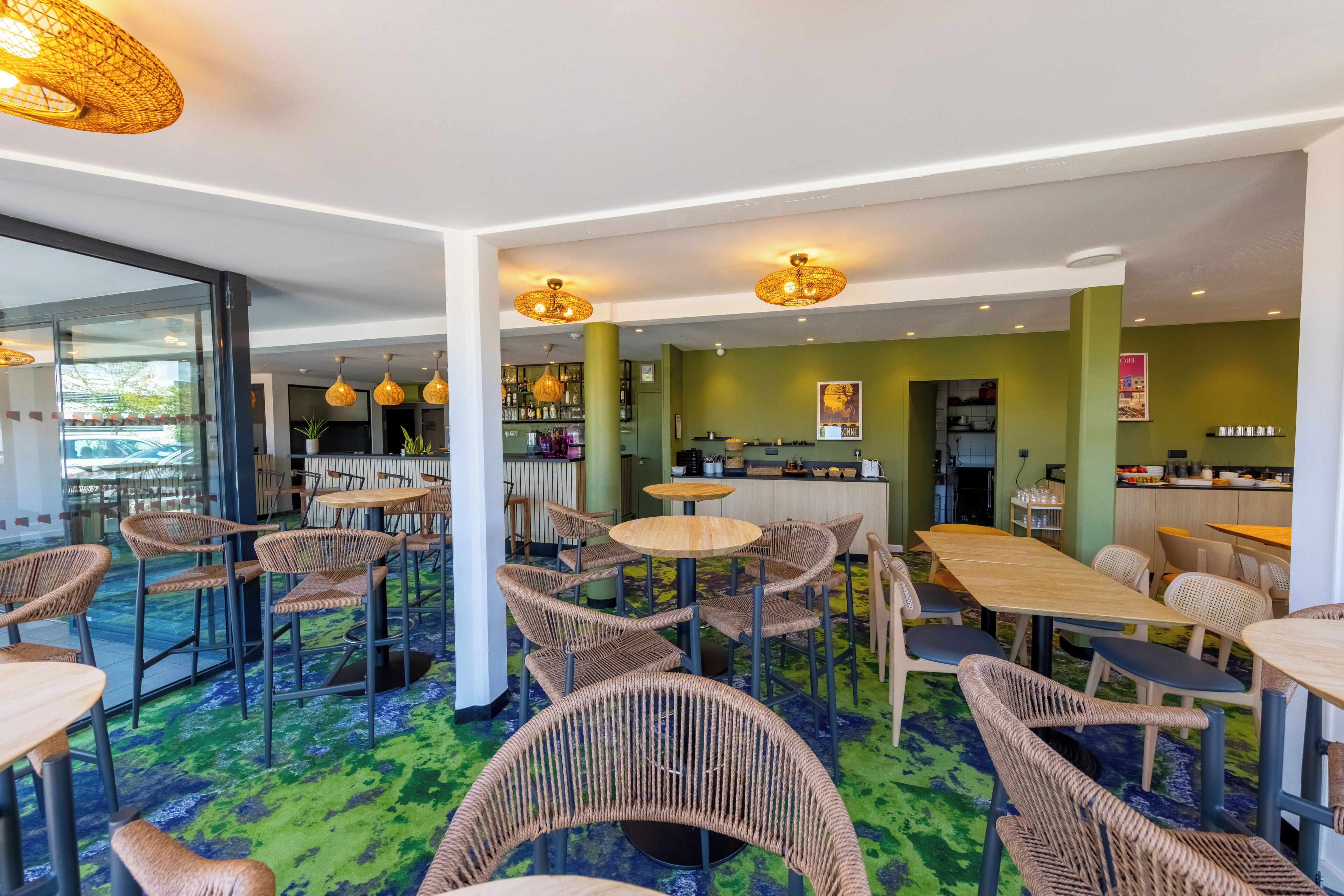 A modern restaurant with wicker chairs and tables, a bar area, and a kitchen visible in the background.