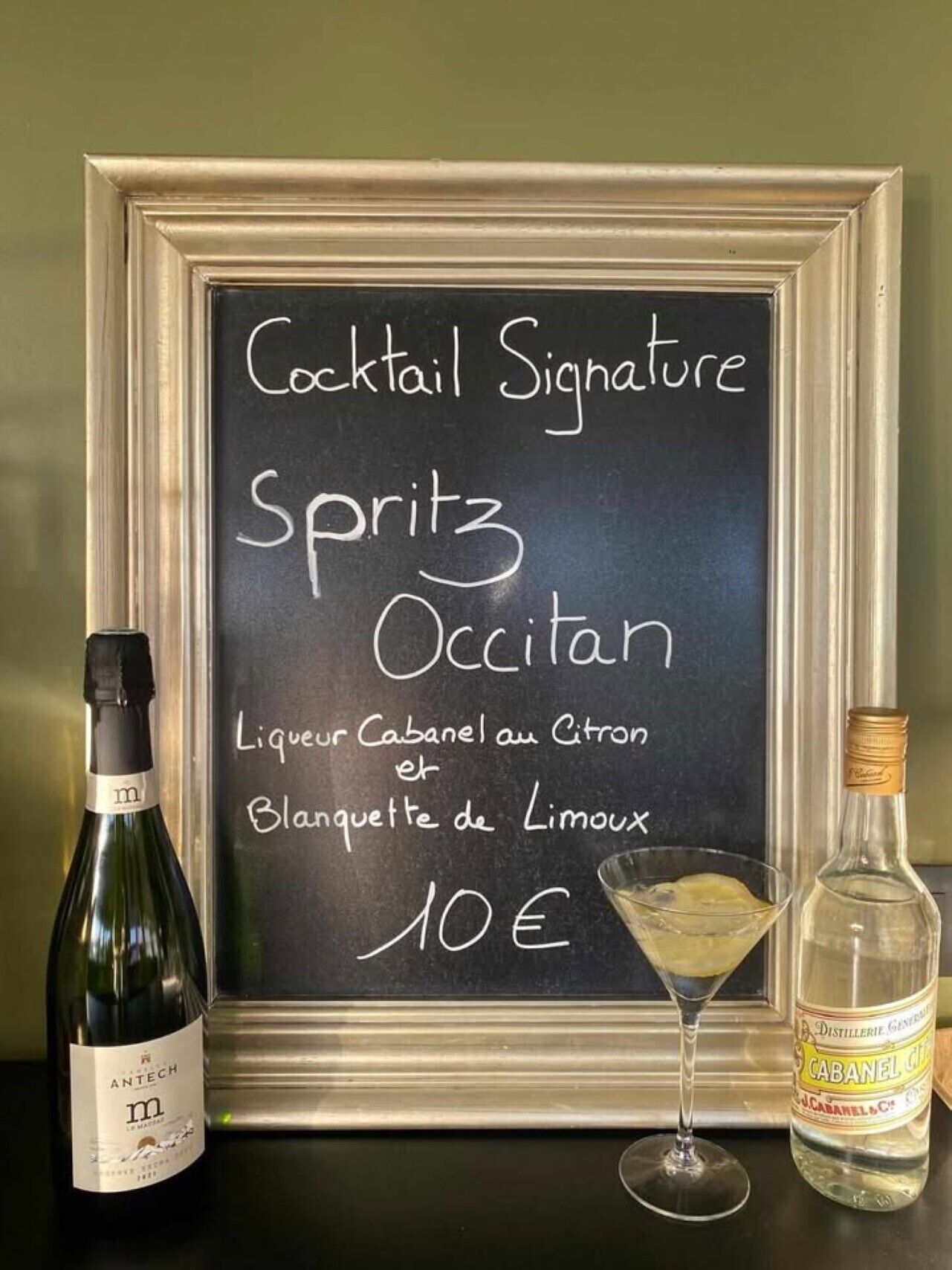 A framed menu board with a cocktail recipe and prices, a bottle of wine, and a martini glass.