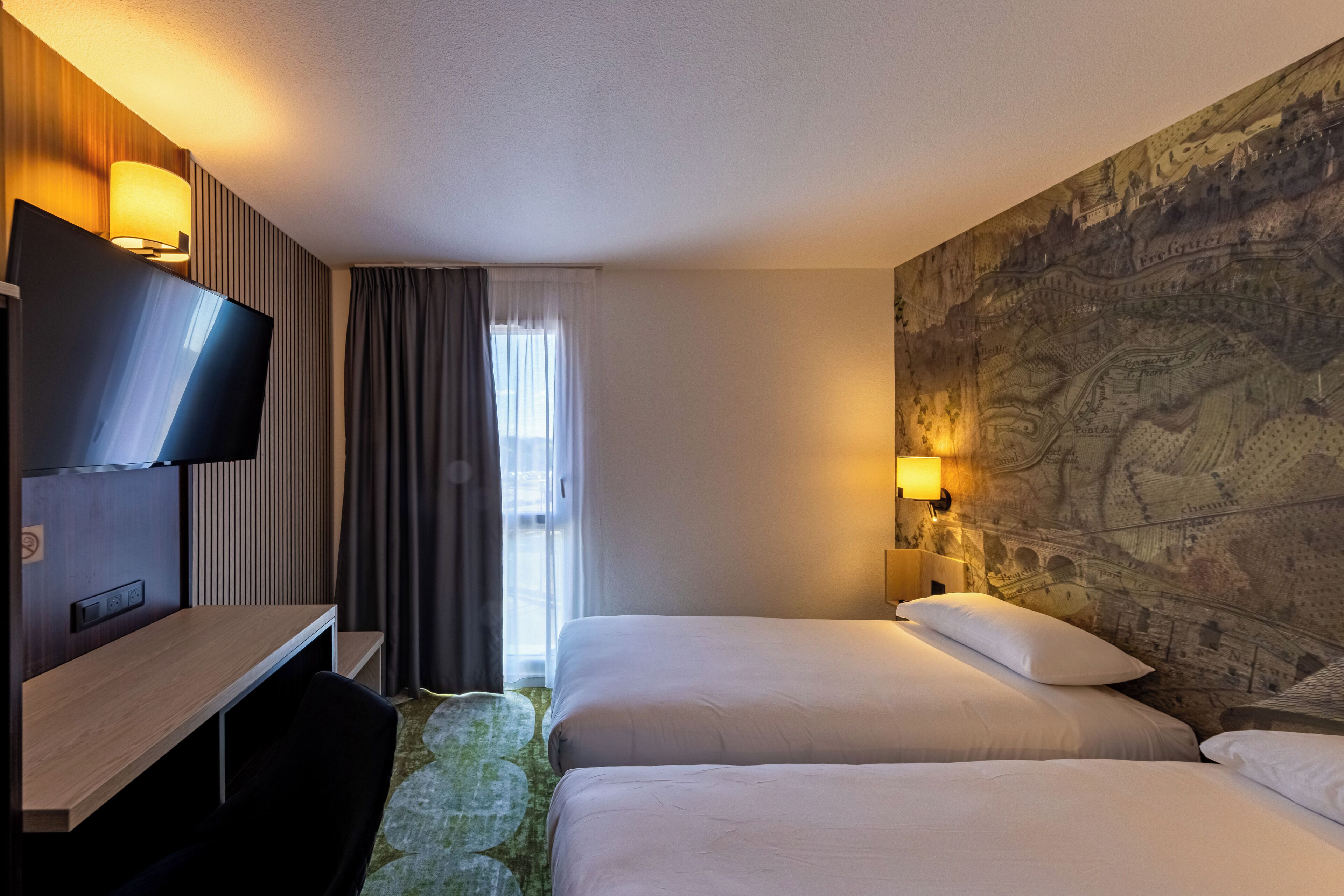 A hotel room with two beds, a desk, a TV, and a wall with a map design.