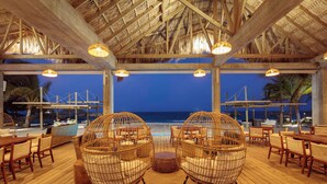An outdoor lounge area with wooden flooring, wicker chairs, and a view of the ocean at dusk. - Indura Beach & Golf Resort, Curio Collection by Hilton (Tela)
