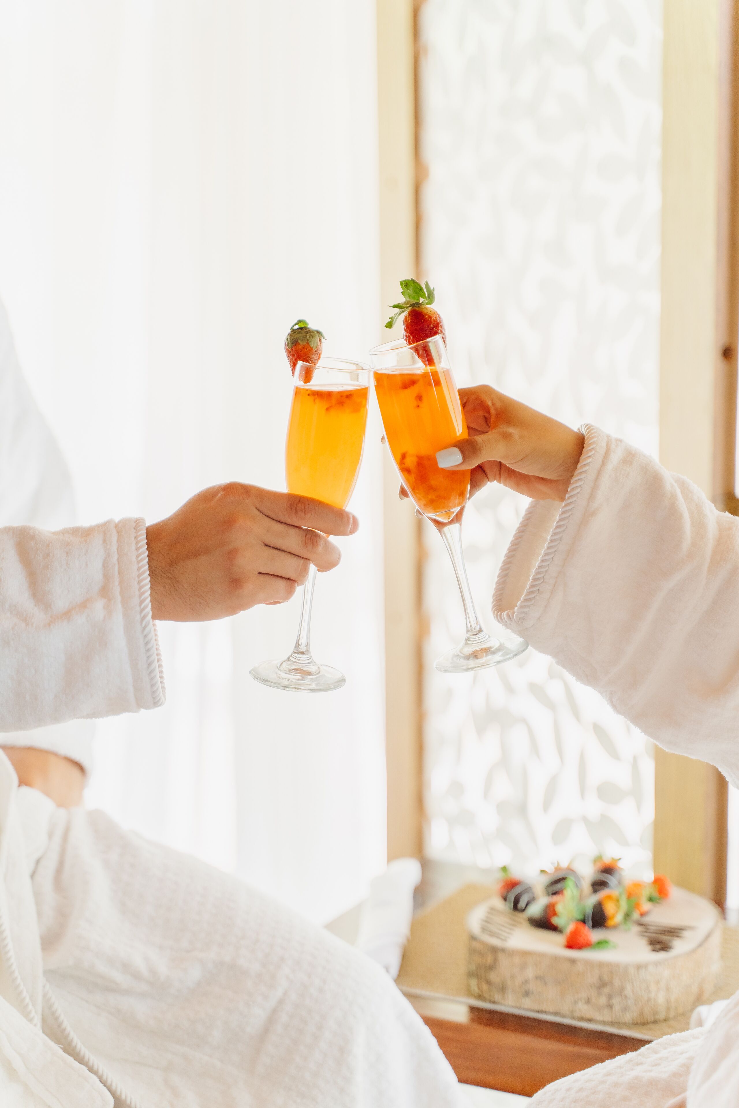 Dos personas sosteniendo copas de champán con fresas, en una habitación luminosa con una mesa blanca y un pastel.