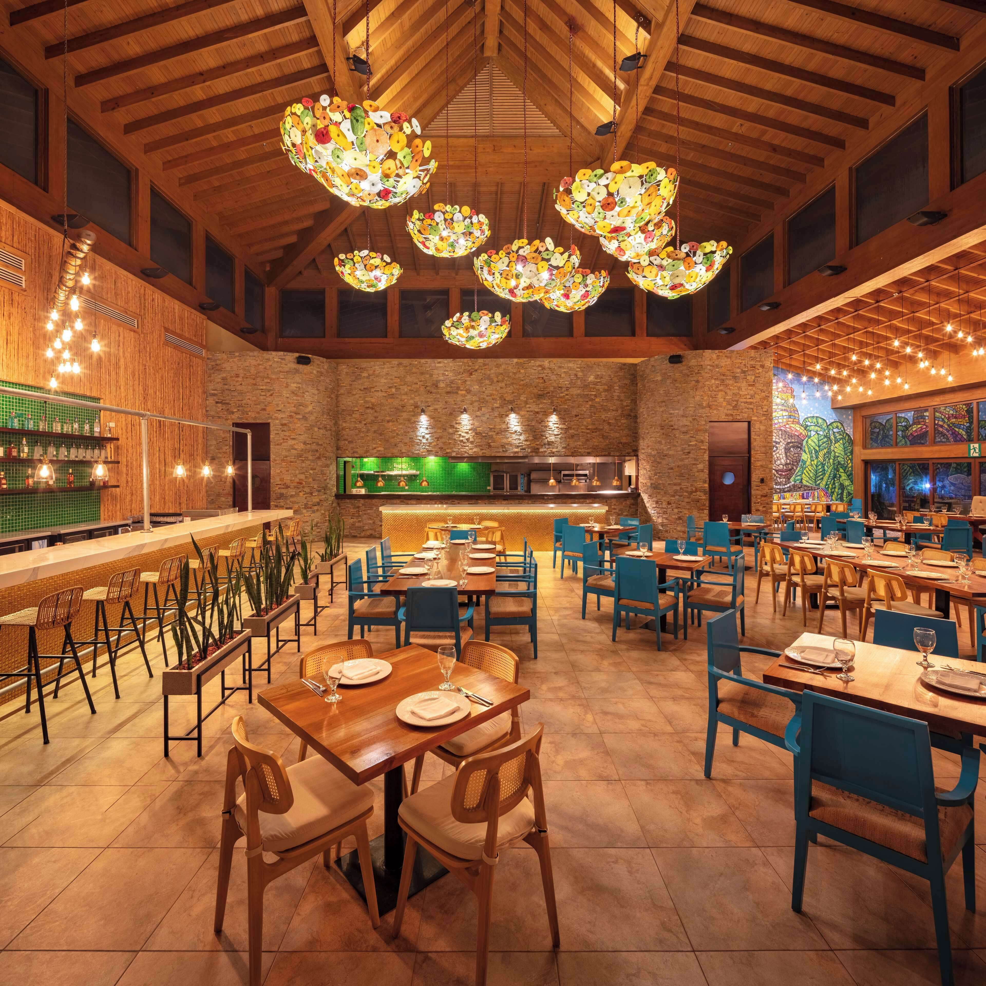 Un restaurant spacieux avec des poutres en bois au plafond, des lustres décorés de motifs de fruits et un bar illuminé de lumière verte.