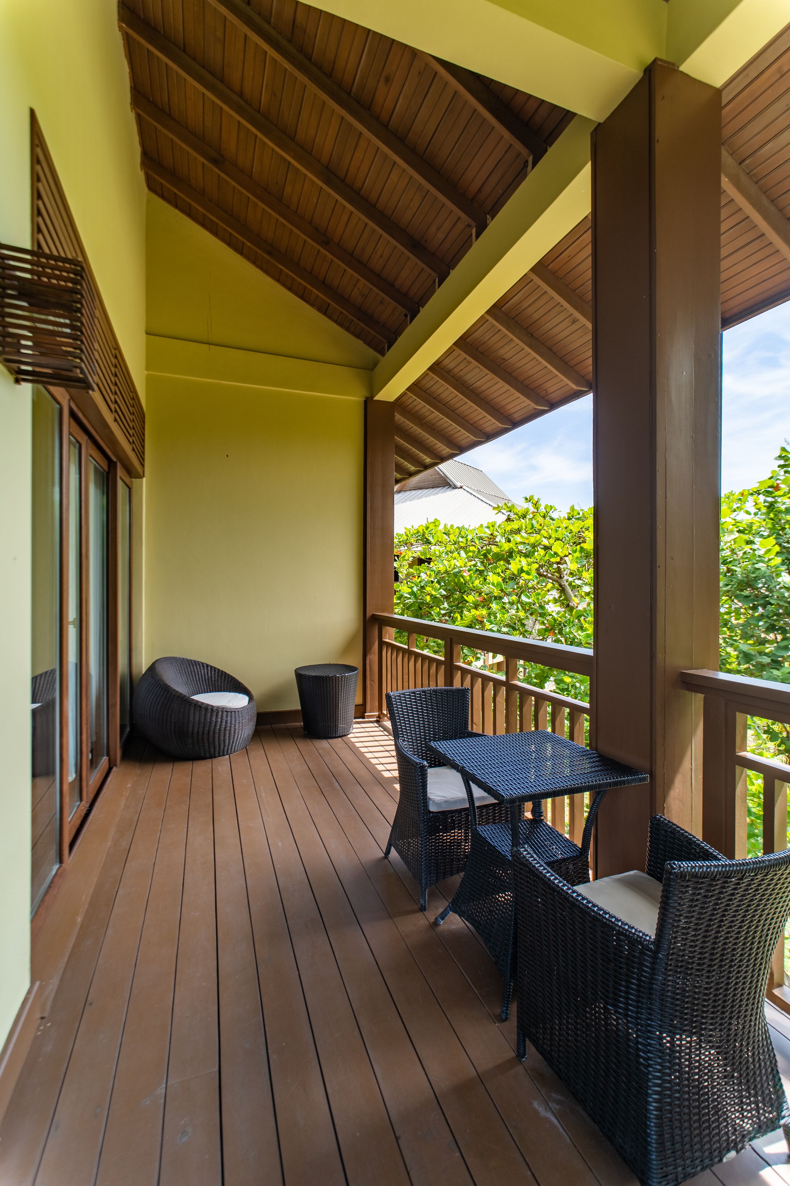 A covered outdoor area with wooden flooring, wicker furniture, and a view of greenery.