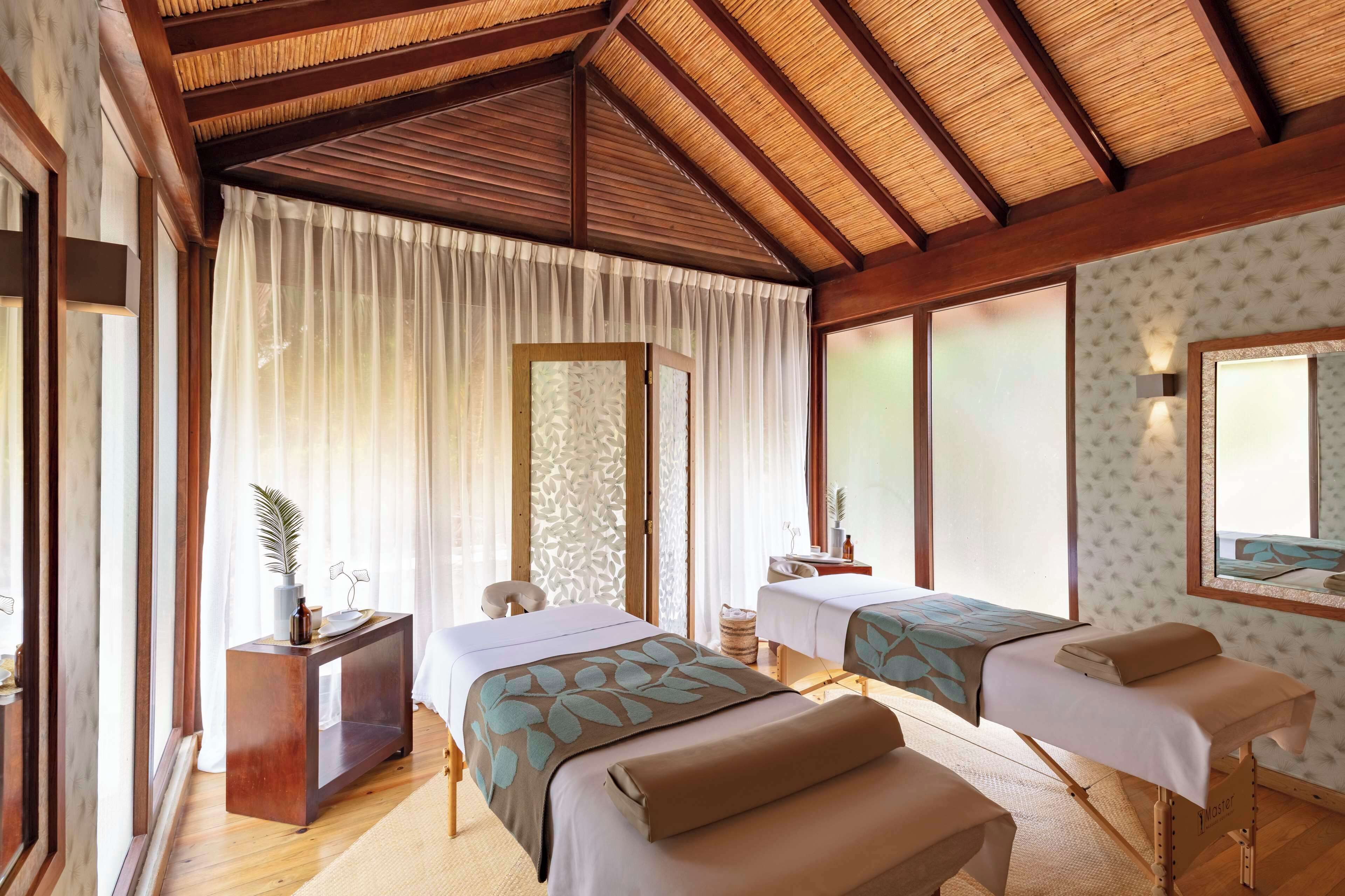 A room with two beds, a small desk, a mirror, and a window with sheer curtains.