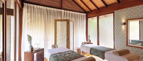 A room with two beds, a small desk, a mirror, and a window with sheer curtains.
