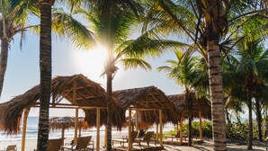 A beach with thatched-roof structures, lounge chairs, and palm trees. - Indura Beach & Golf Resort, Curio Collection by Hilton (Tela)