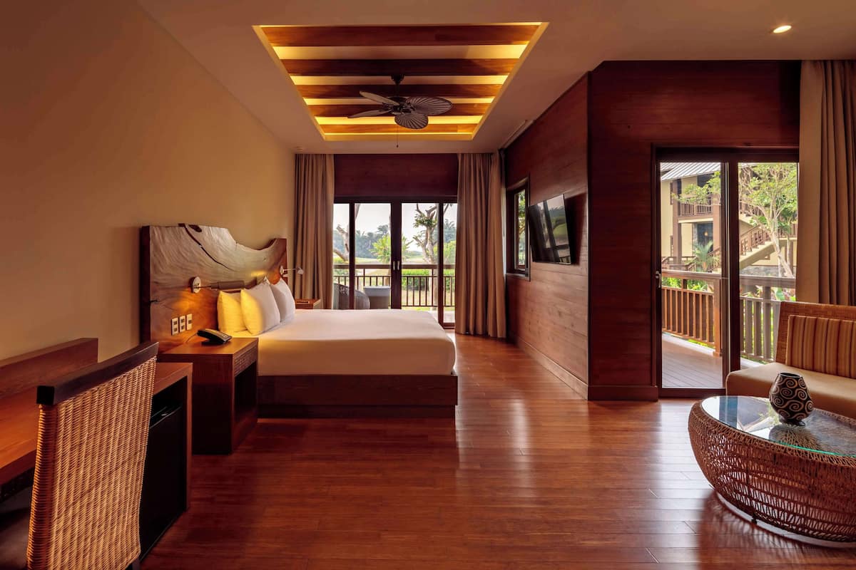 A hotel room with a bed, a desk with a lamp, a chair, a round wicker coffee table, and a balcony with a view of the outdoors.