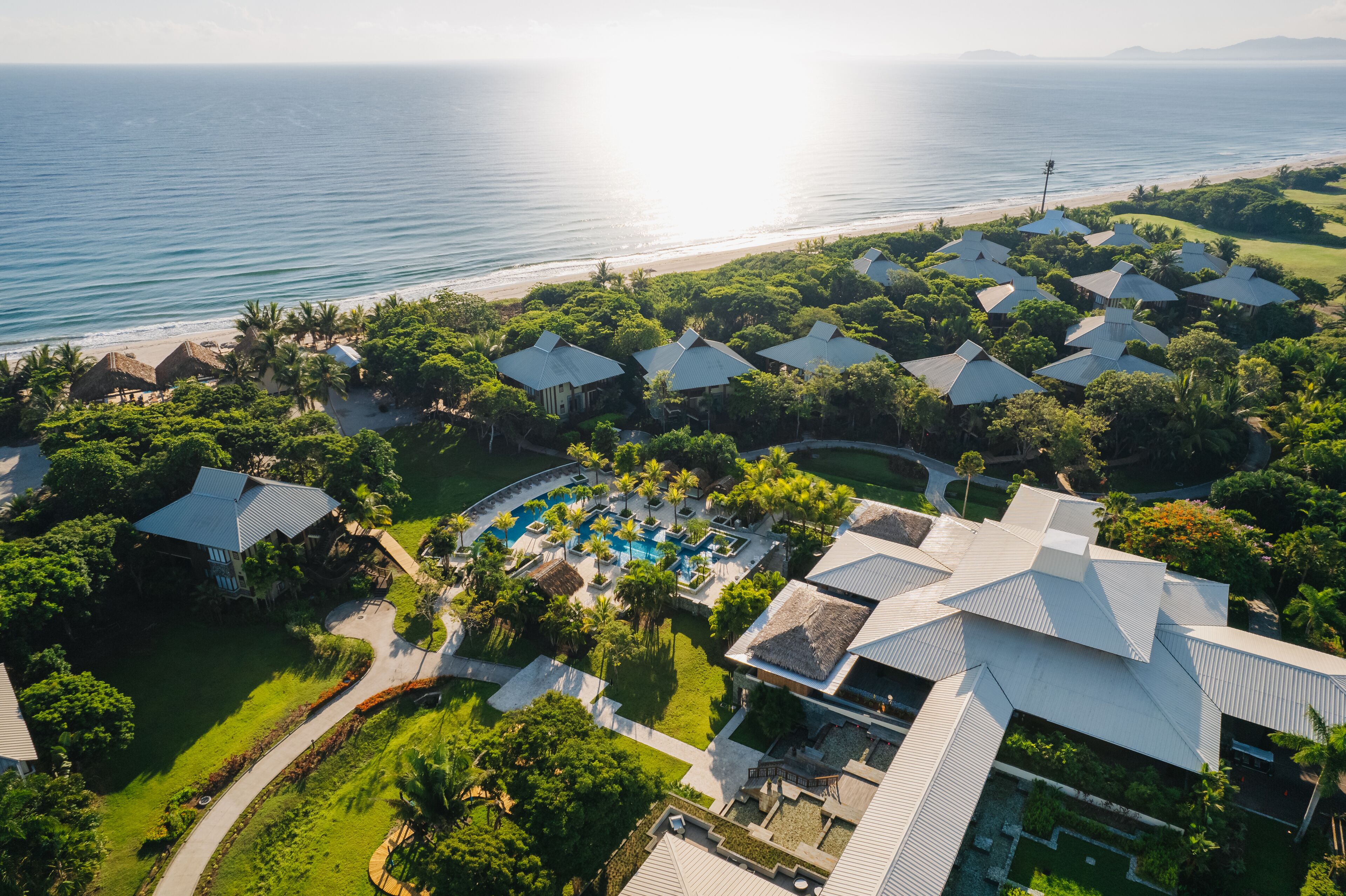 Une vue aérienne d’un complexe hôtelier comprenant plusieurs bâtiments de style bungalow, un espace piscine central et une plage en arrière-plan.
