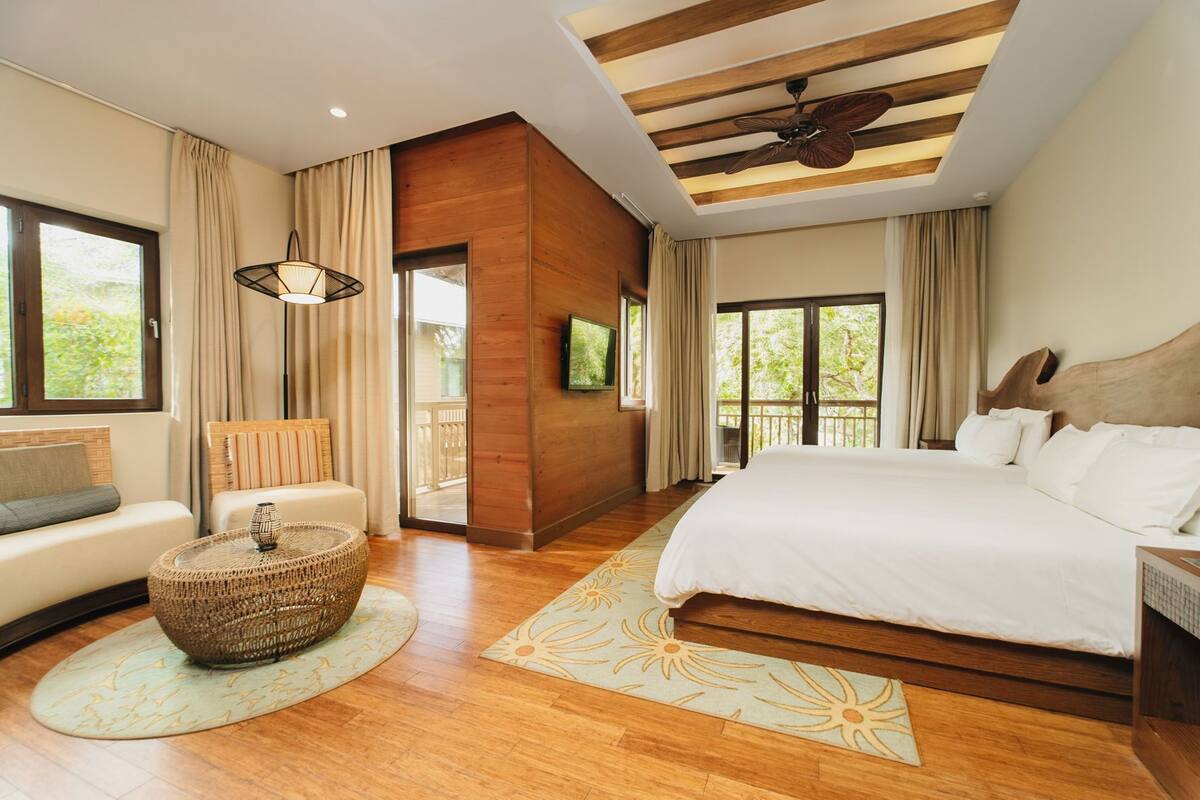 A modern hotel room with a large bed, a sofa, a round coffee table, and a balcony with a view.
