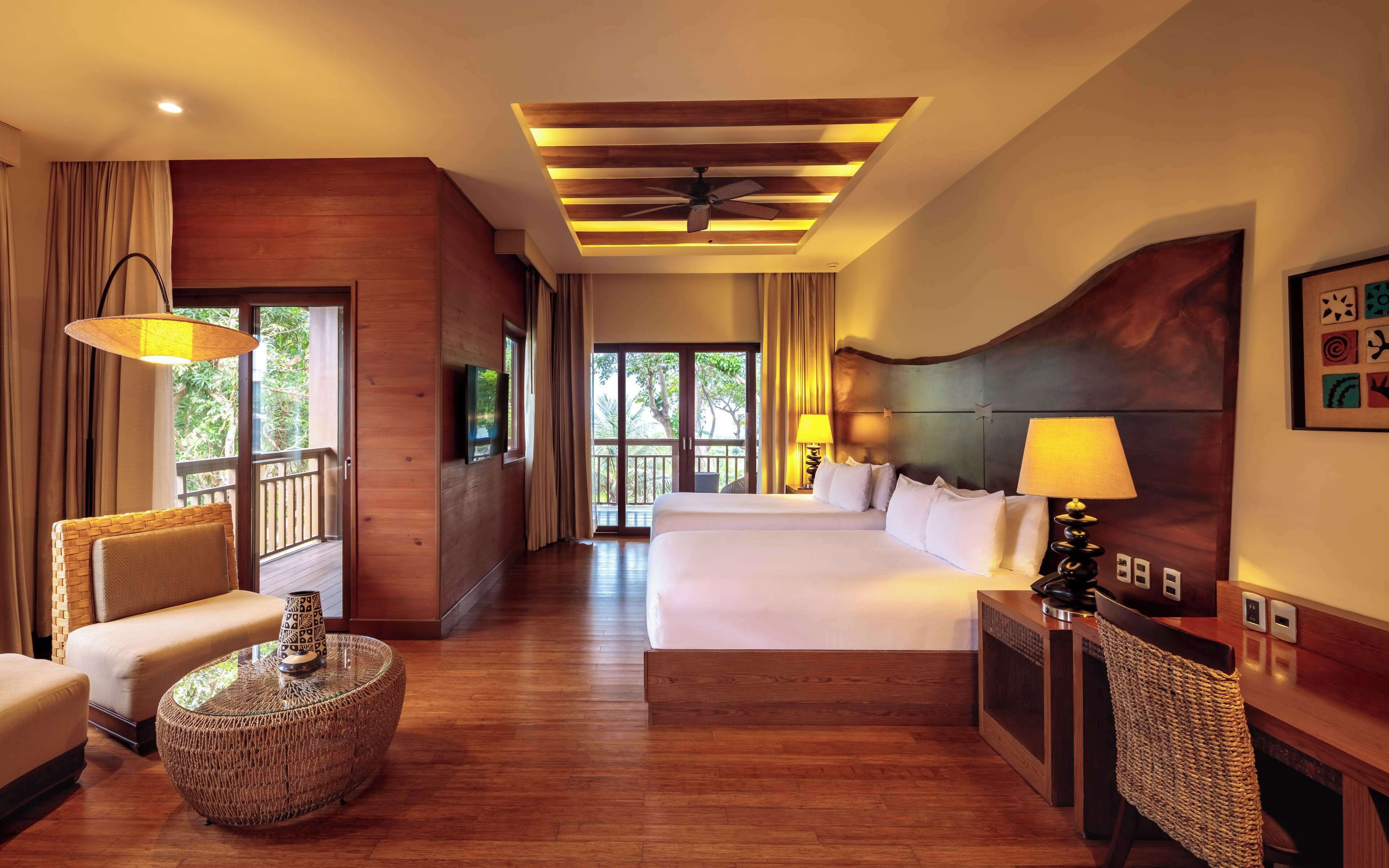 A hotel room with a large bed, a sofa, a round wicker coffee table, a ceiling fan, and a view of the outdoors.