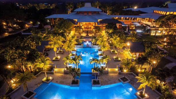 A large, illuminated swimming pool surrounded by palm trees and lounge chairs, with a building in the background. - Indura Beach & Golf Resort, Curio Collection by Hilton (Tela)