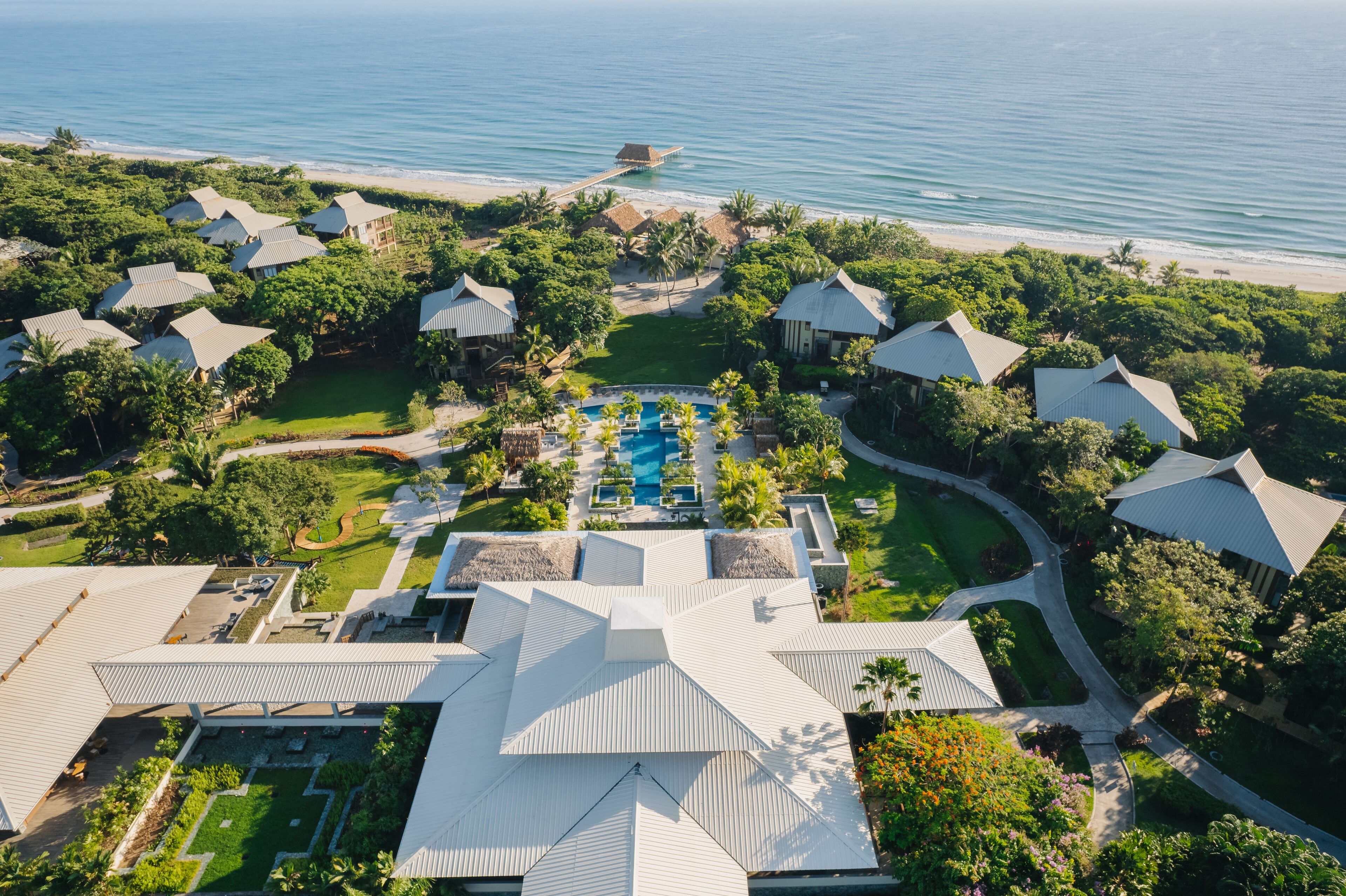 Vista aérea de un complejo turístico con varias cabañas, una piscina central y una playa al fondo.