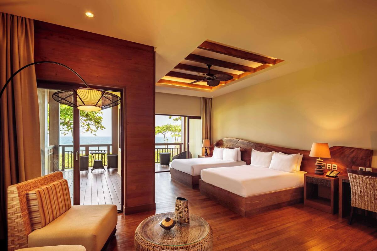 A hotel room with two beds, a sofa, a round coffee table, and a balcony with a view of the sea.