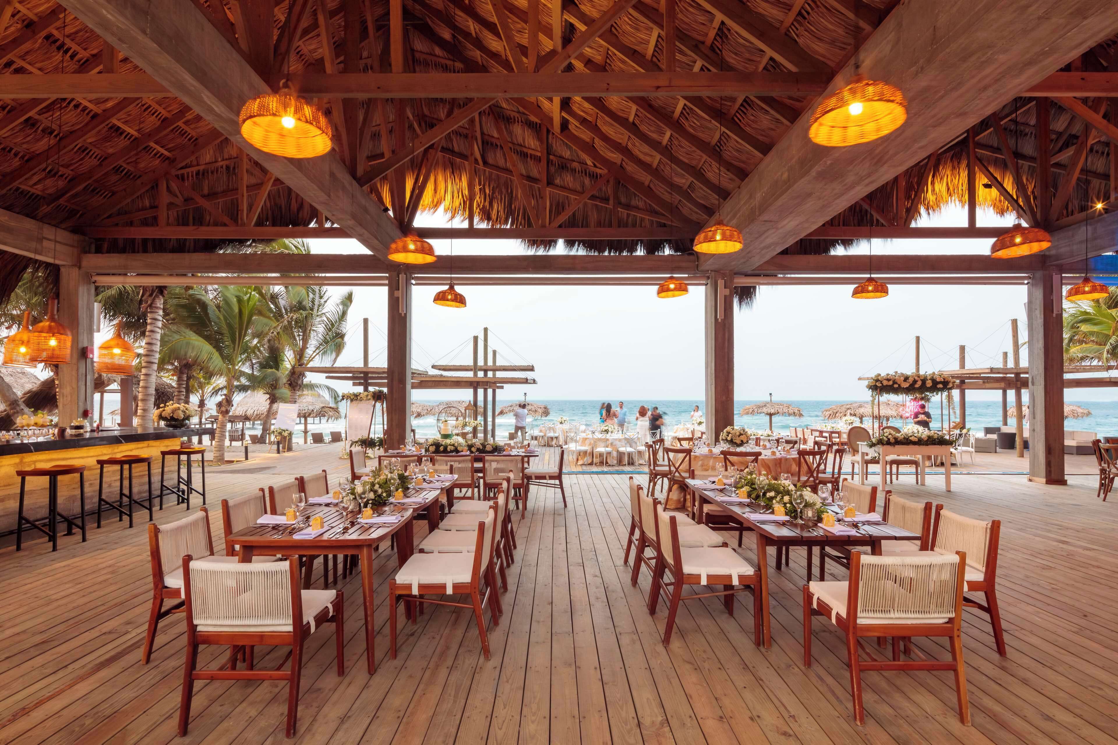 An outdoor dining area with wooden tables and chairs, a thatched roof, and a view of the beach.