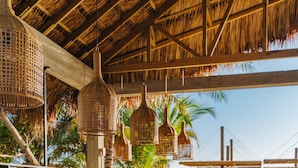 A wooden deck with bar stools and hanging wicker lamps, overlooking a pool and palm trees. - Indura Beach & Golf Resort, Curio Collection by Hilton (Tela)