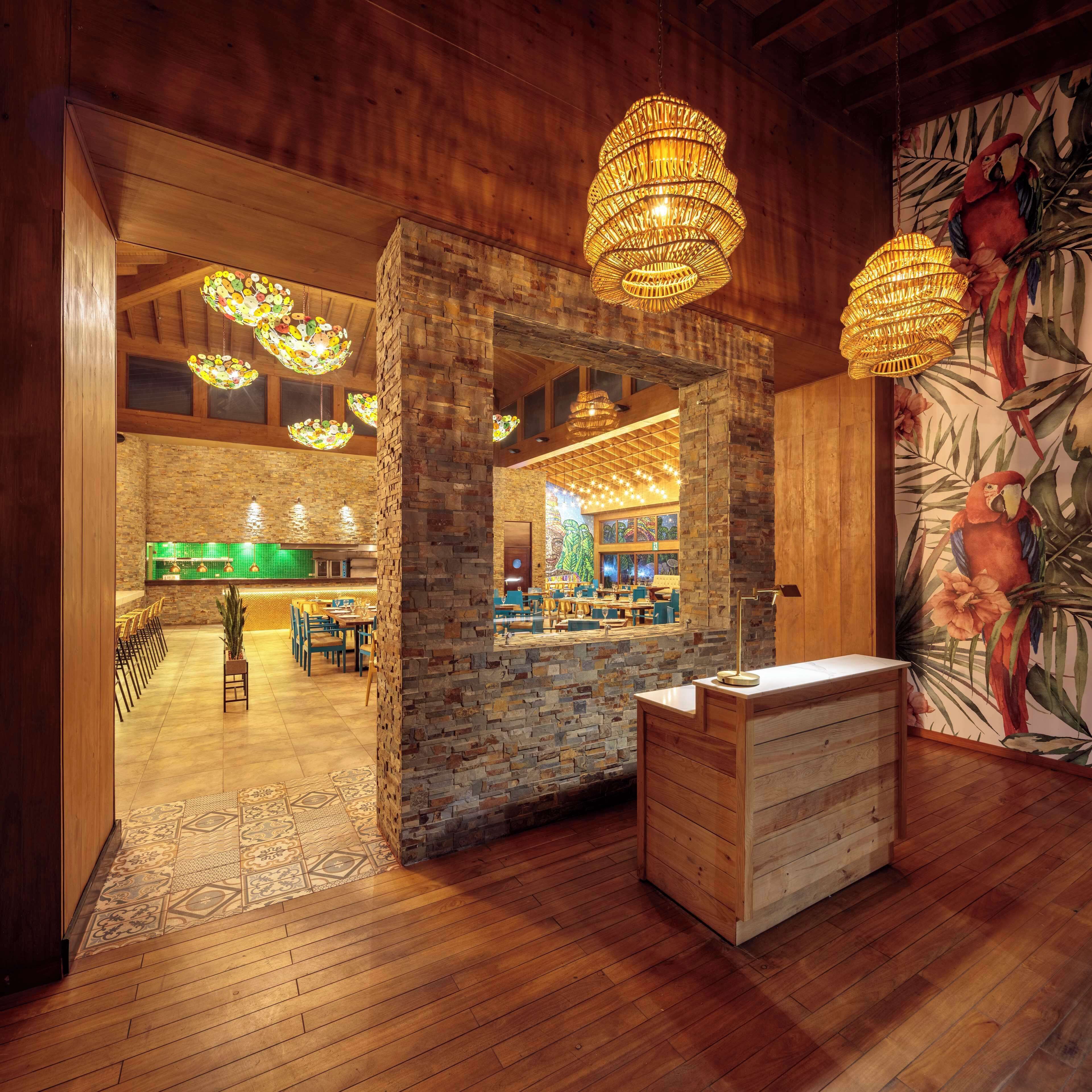 Een restaurant interieur met houten wanden, een accentmuur van steen en een tropische muurschildering. Er zijn hangende lampen en een receptiebalie.
