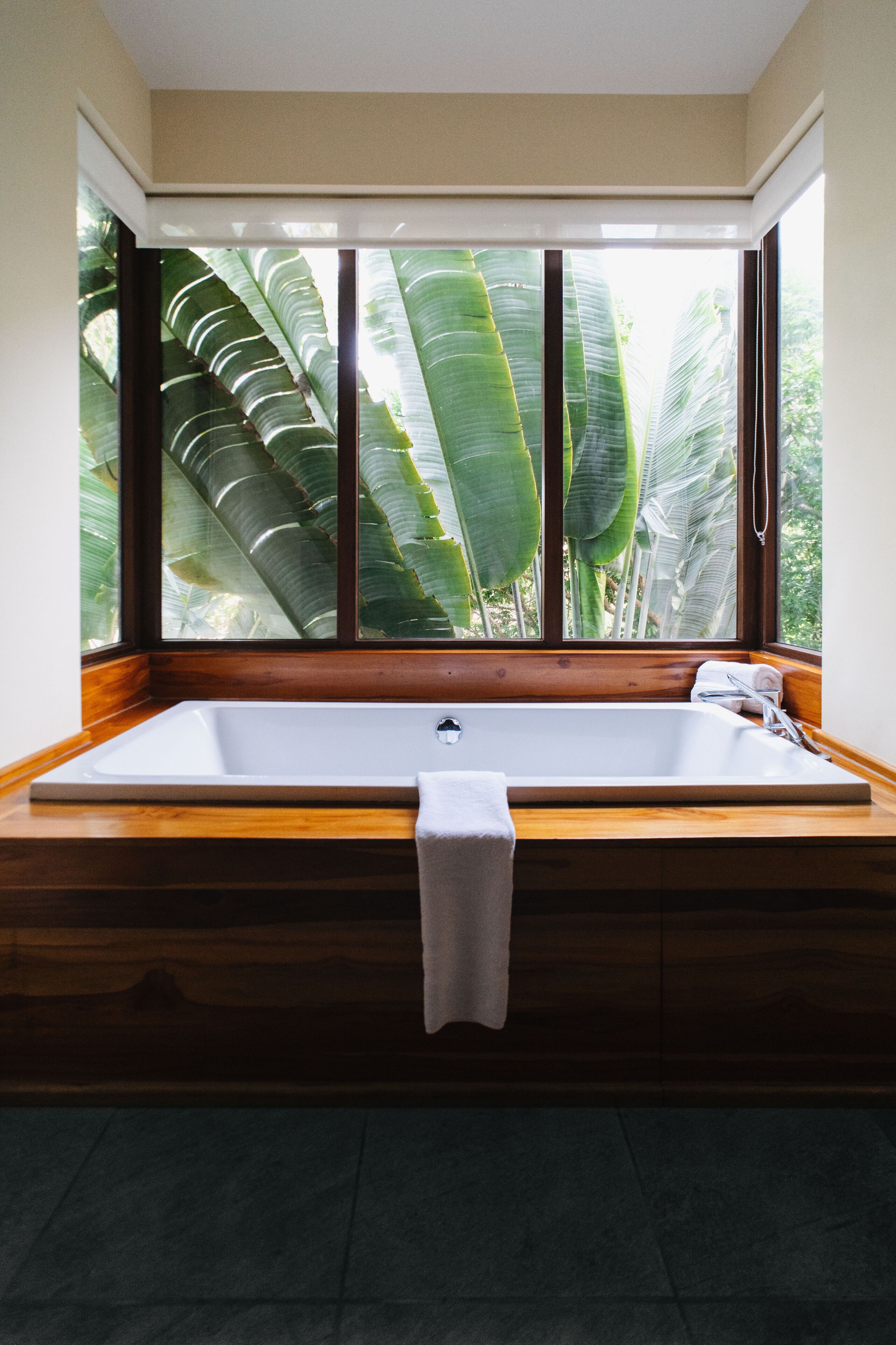 A large bathtub with a wooden base, a white towel, and a window with a view of tropical foliage.