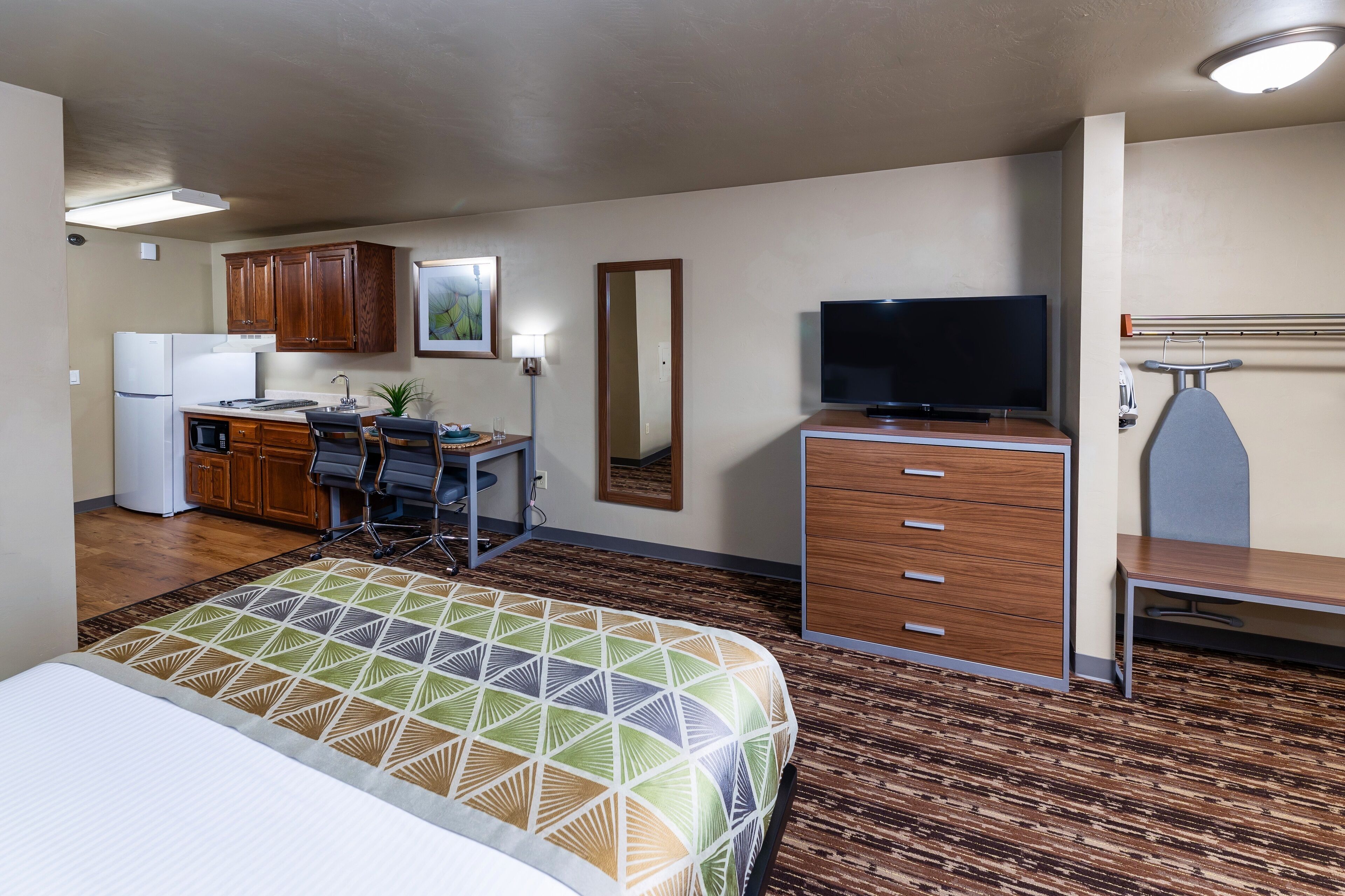 A compact hotel room with a kitchenette, a bed with a patterned bedspread, a television, and a desk with a chair.