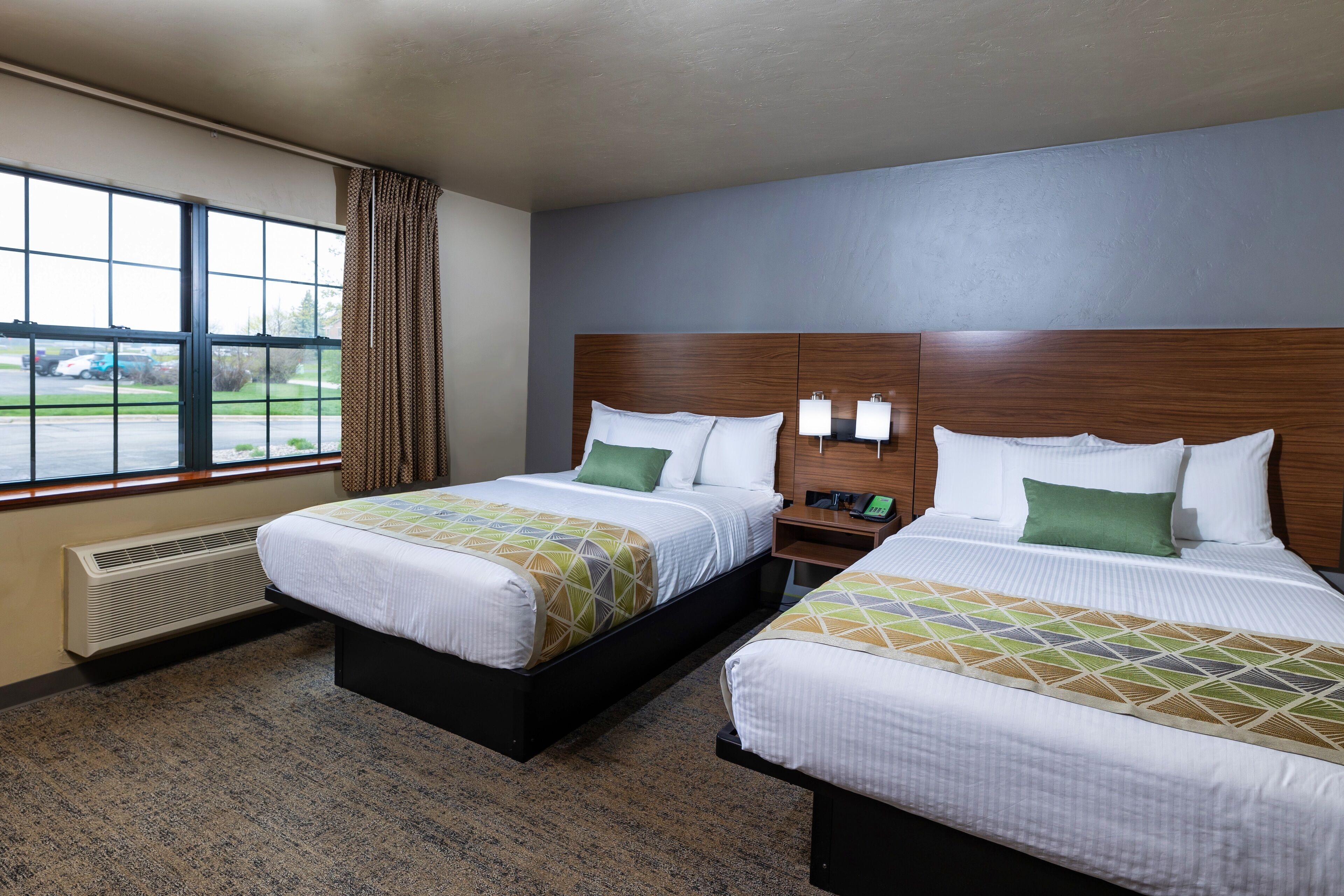 A hotel room with two beds, a large window, a heater, and a nightstand with a lamp.