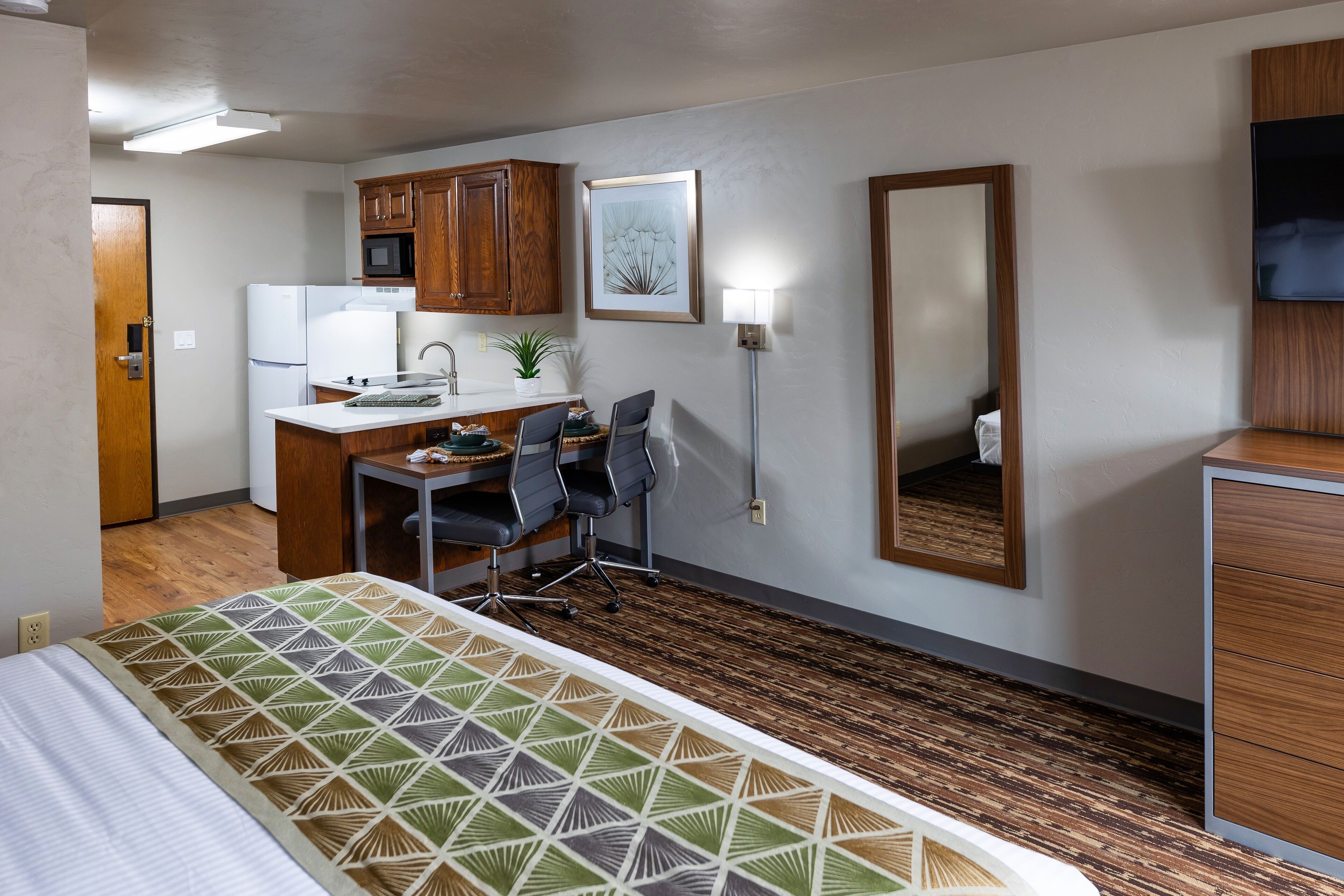 A compact hotel room with a bed, a kitchenette, a desk with chairs, a mirror, and a flat-screen TV.