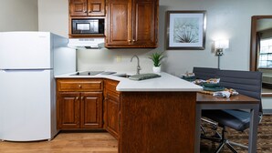 A compact kitchen with wooden cabinets, a microwave, a sink, and a dining area with a chair. - Extended Stay Airport (Green Bay)