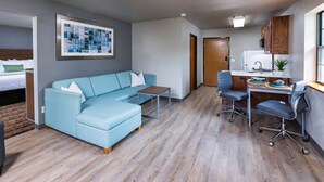 A modern living room with a blue sectional sofa, a small wooden coffee table, and a desk with a chair. There is a kitchen area with a refrigerator and a sink. - Extended Stay Airport (Green Bay)