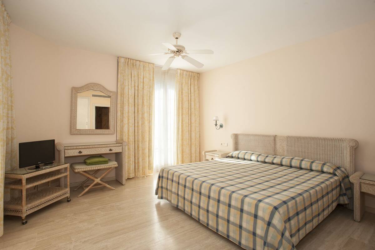 A bedroom with a bed, a desk, a television, a mirror, and a ceiling fan.