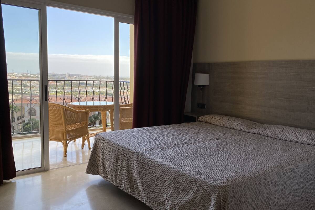 A hotel room with a bed, a balcony with wicker furniture, and a city view.