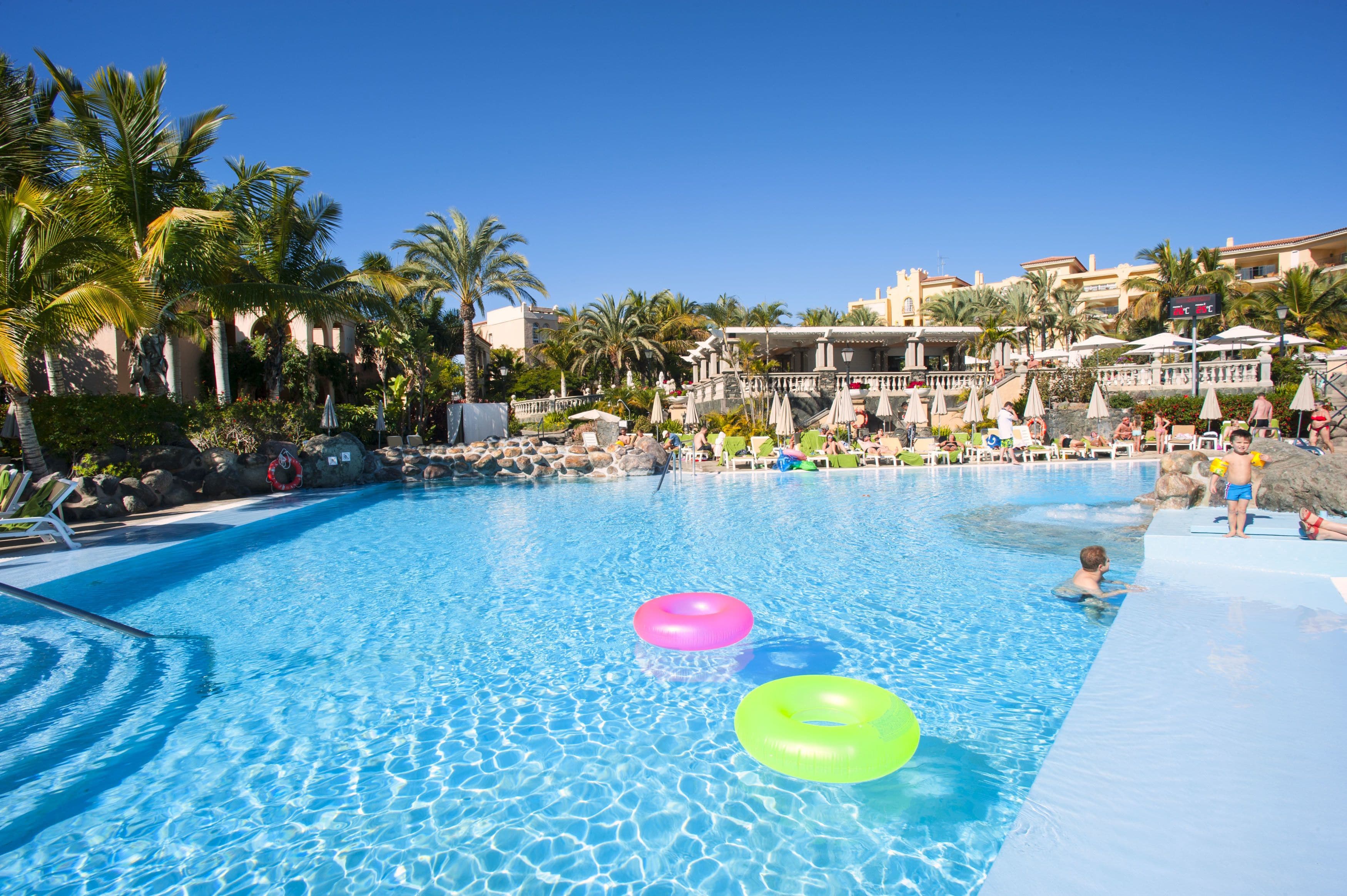 A swimming pool with clear blue water, surrounded by palm trees and a resort building with balconies.