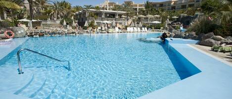 A large outdoor swimming pool with clear blue water, surrounded by lounge chairs and umbrellas, with a multi-story building in the background.