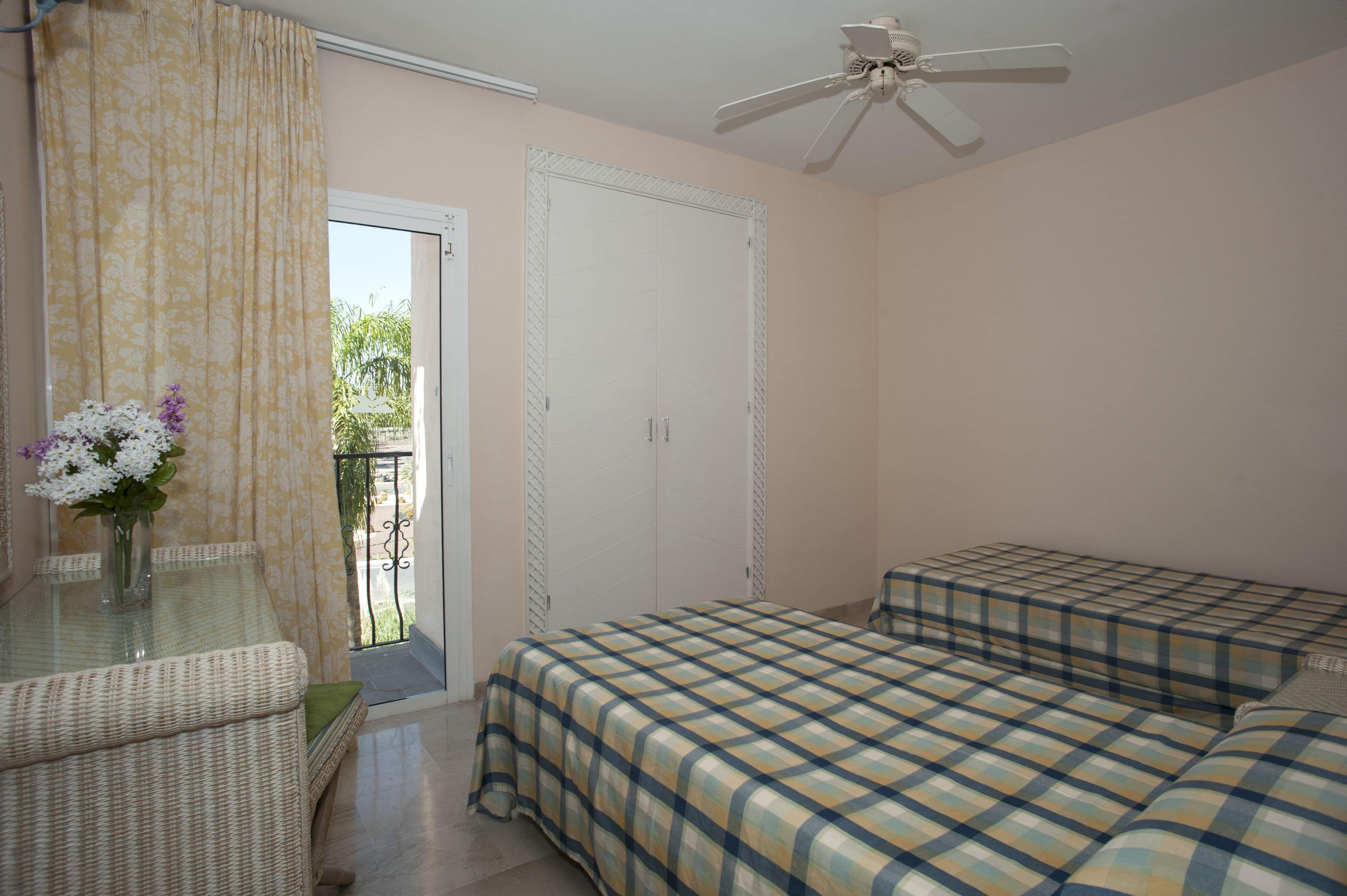 A bedroom with a bed, a small table with a vase of flowers, a ceiling fan, and a door leading to a balcony.