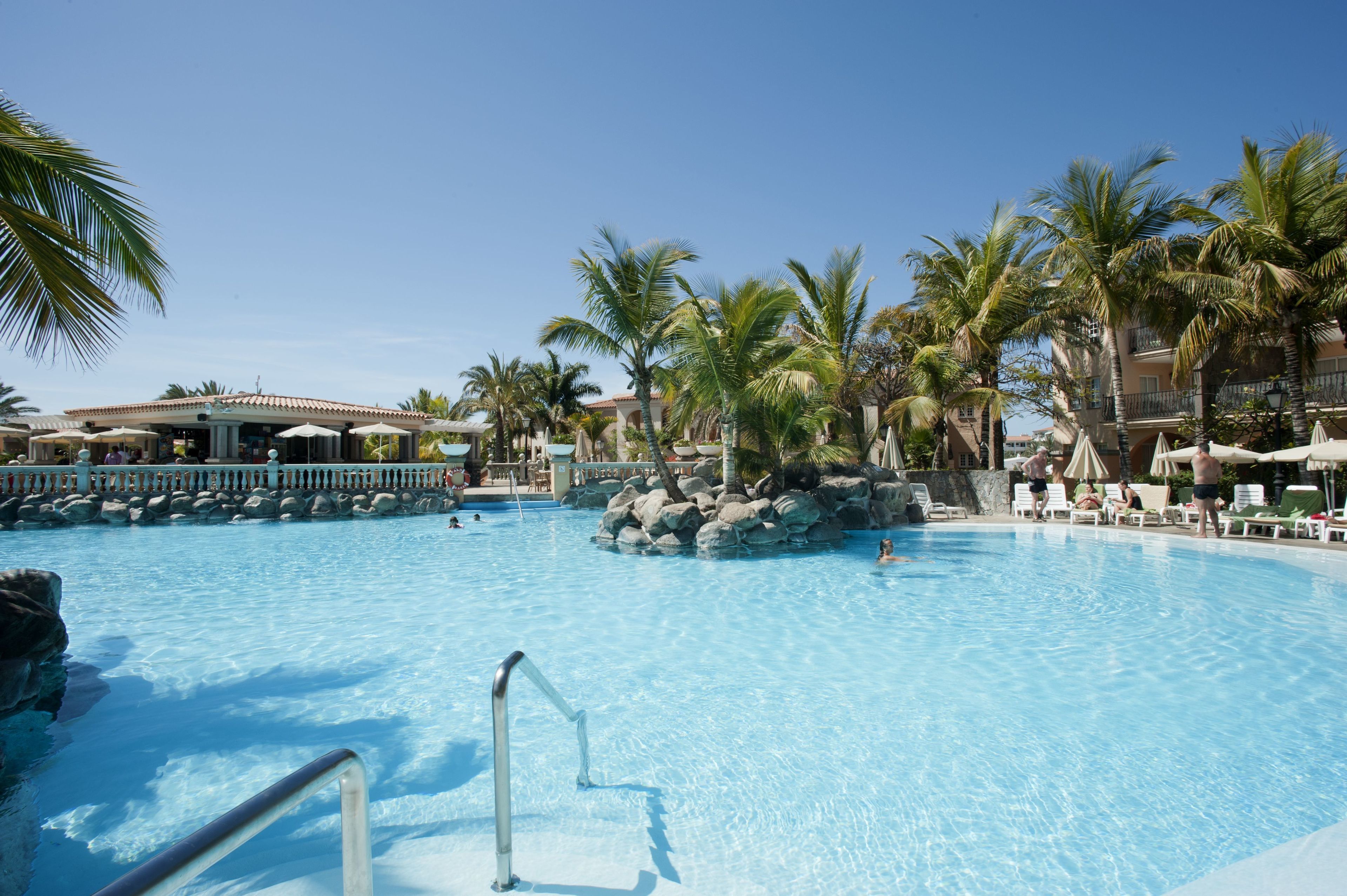 A clear blue pool surrounded by palm trees and lounge chairs, with a building in the background.