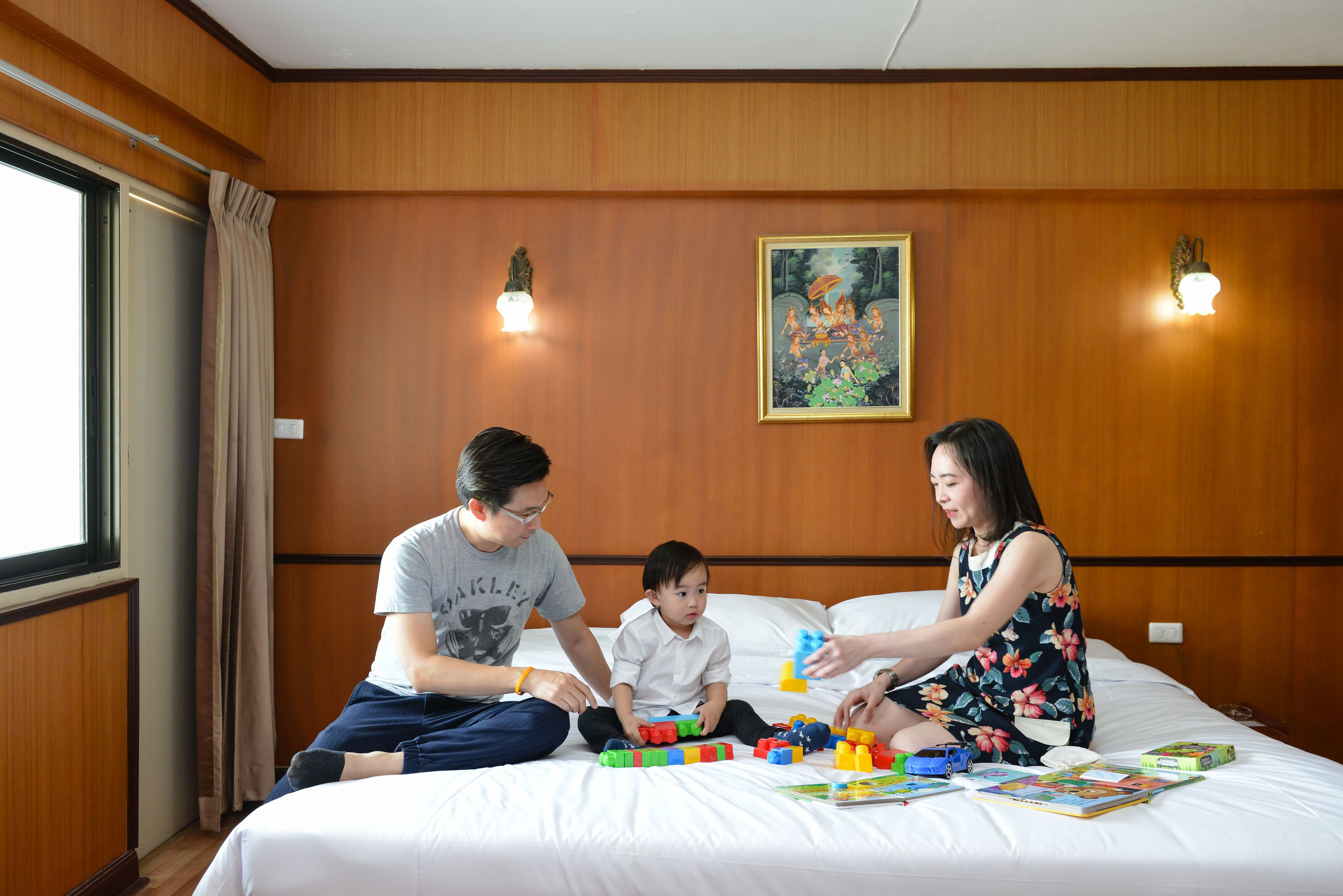 A hotel room with wooden paneling, a bed with white linens, a window with curtains, and a painting on the wall.