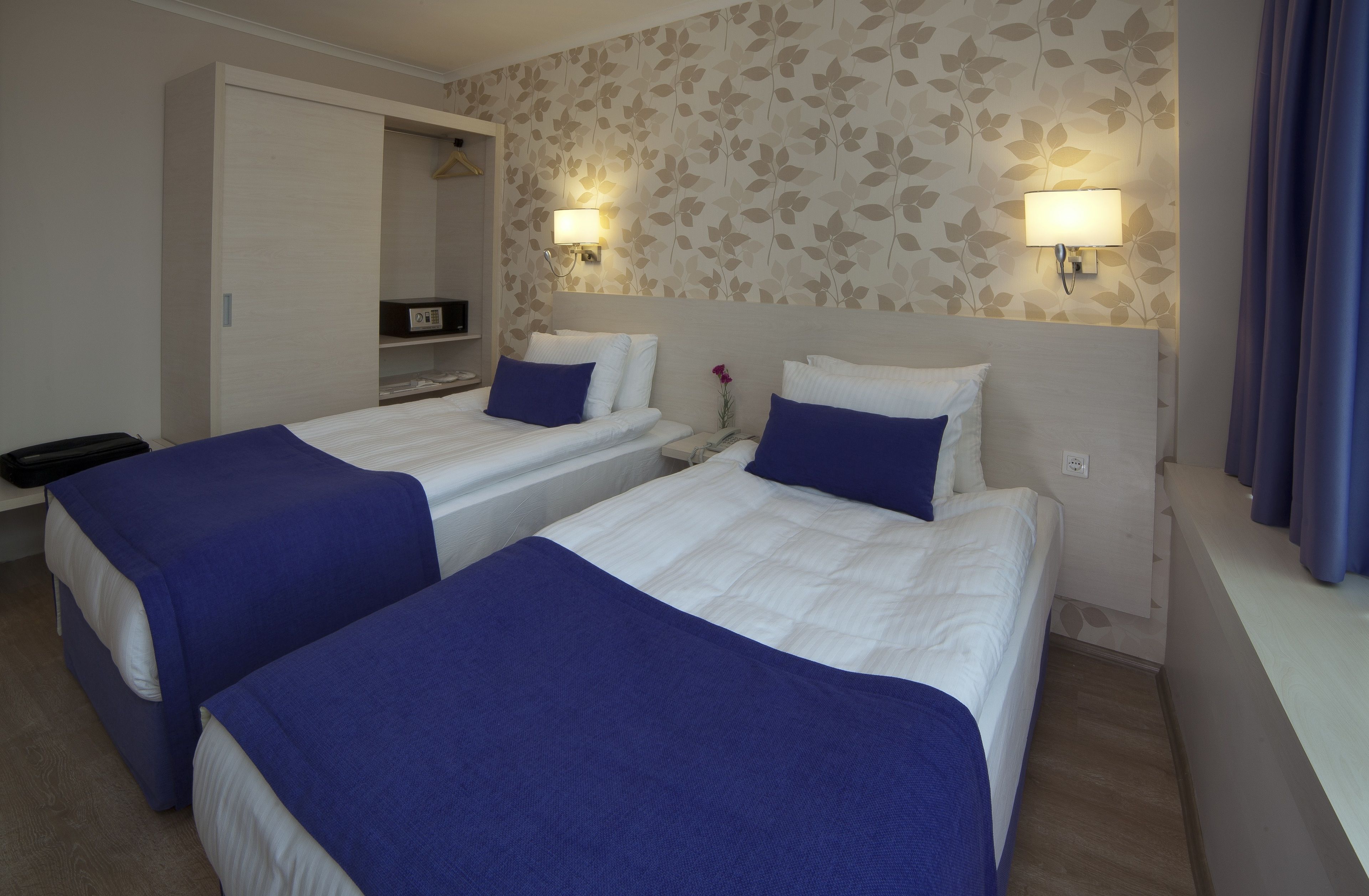 A hotel room with two beds, each with a blue bedspread and white pillows, a wardrobe, a desk with a lamp, and a floral wallpaper.