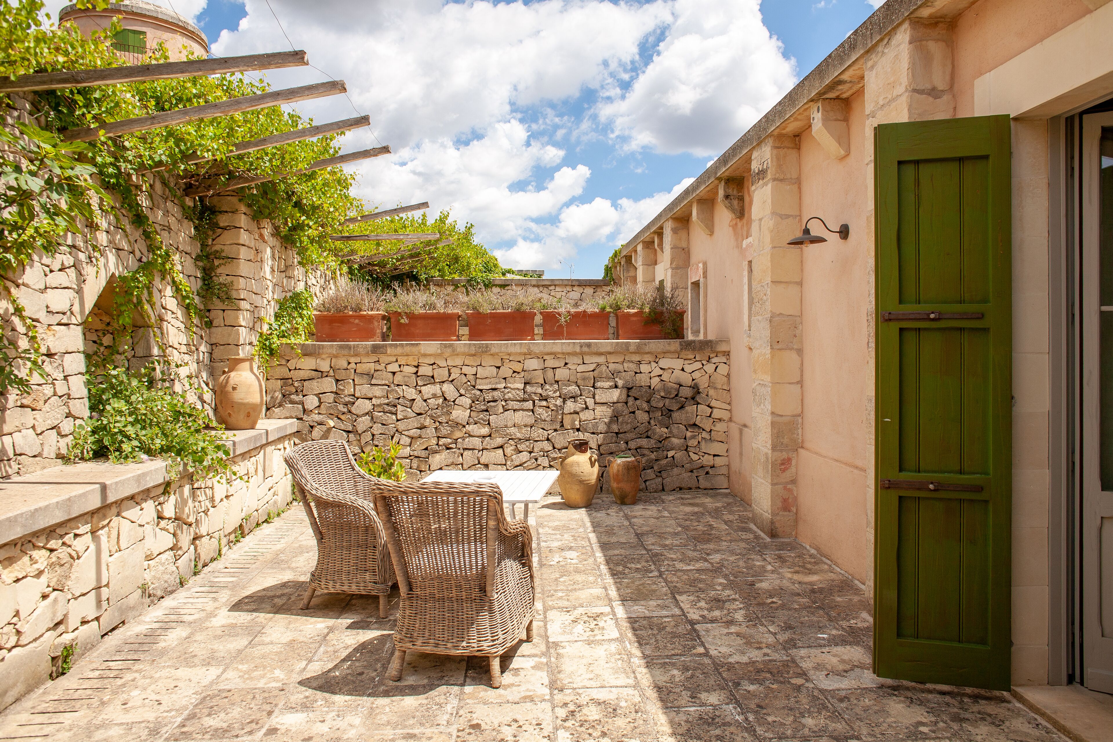 A stone-walled patio with wicker furniture, a green door, and a stone wall with climbing plants.