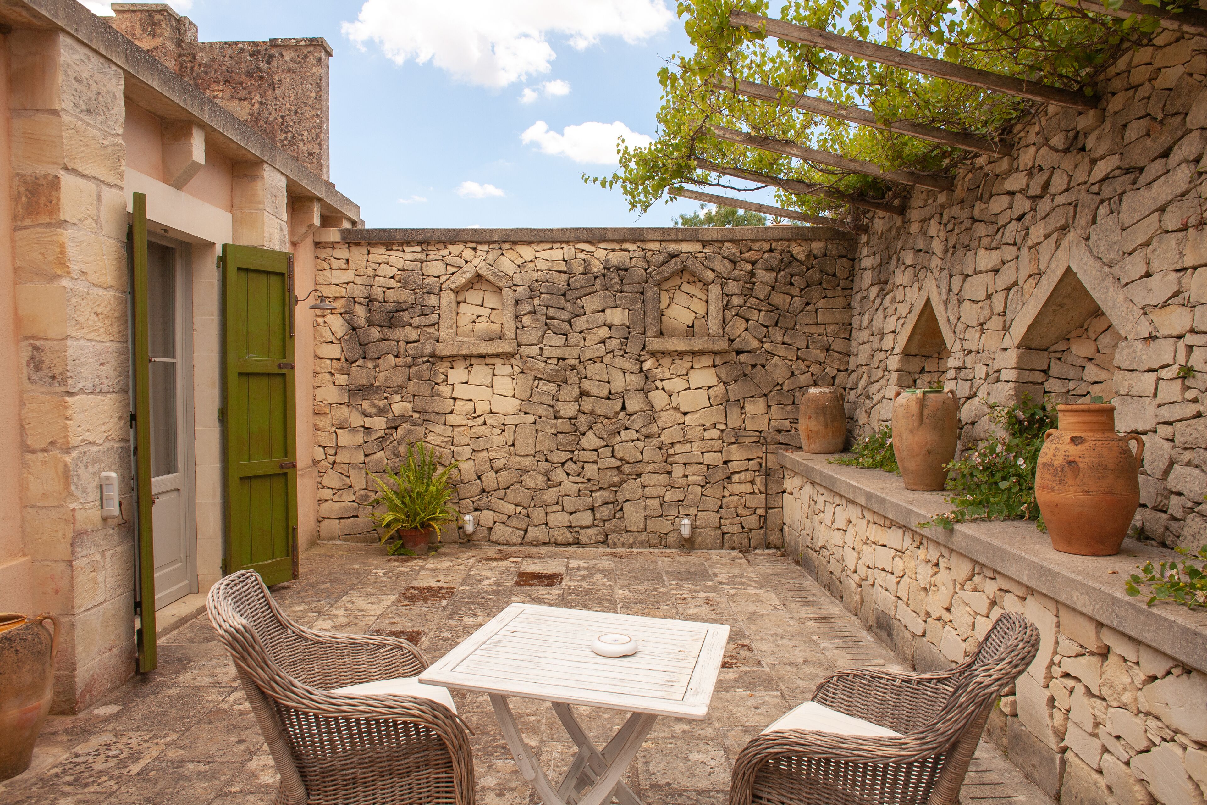 A stone-walled patio with wicker furniture, terracotta pots, and a pergola.