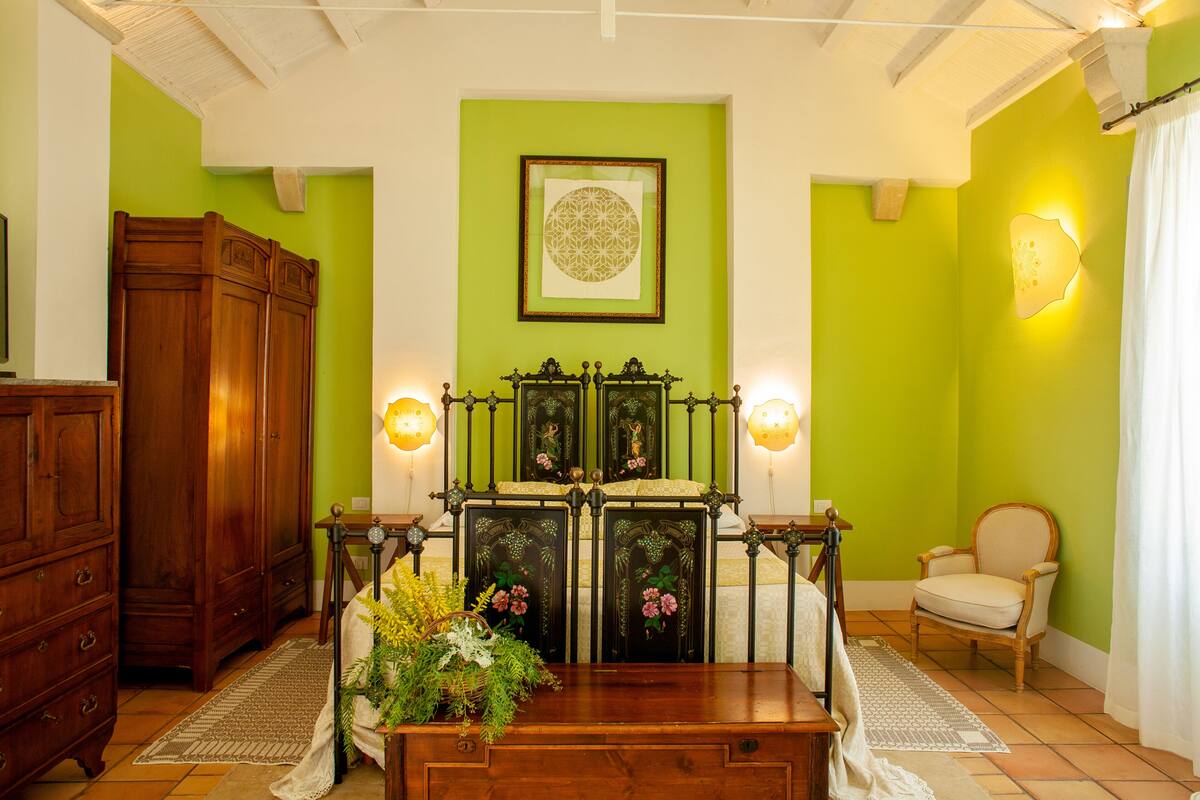 A room with a green wall, a wooden cabinet, a bed with a headboard, a chair, and a framed artwork on the wall.