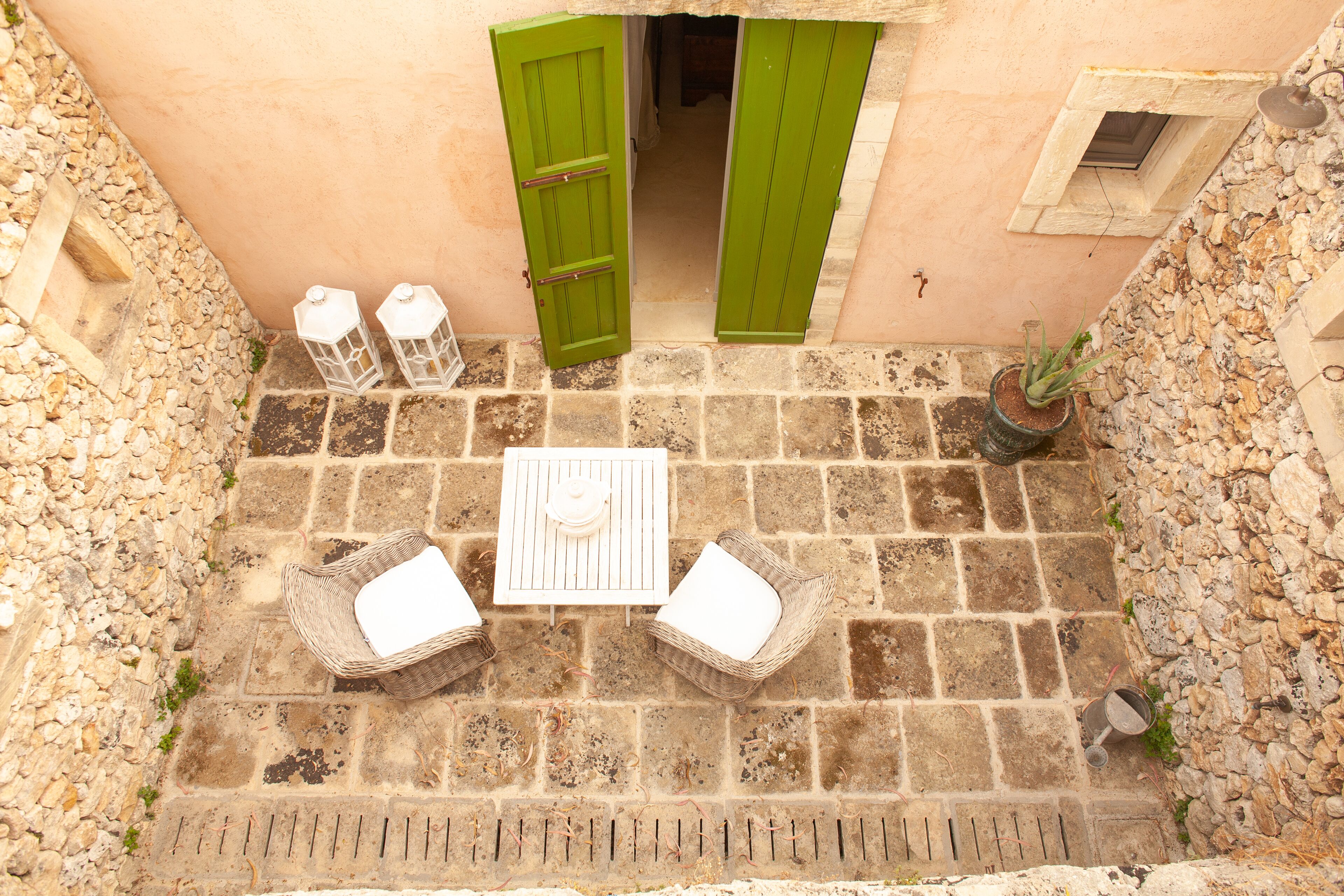 Un patio de piedra con una mesa y dos sillas de mimbre, una puerta verde y una planta en maceta.