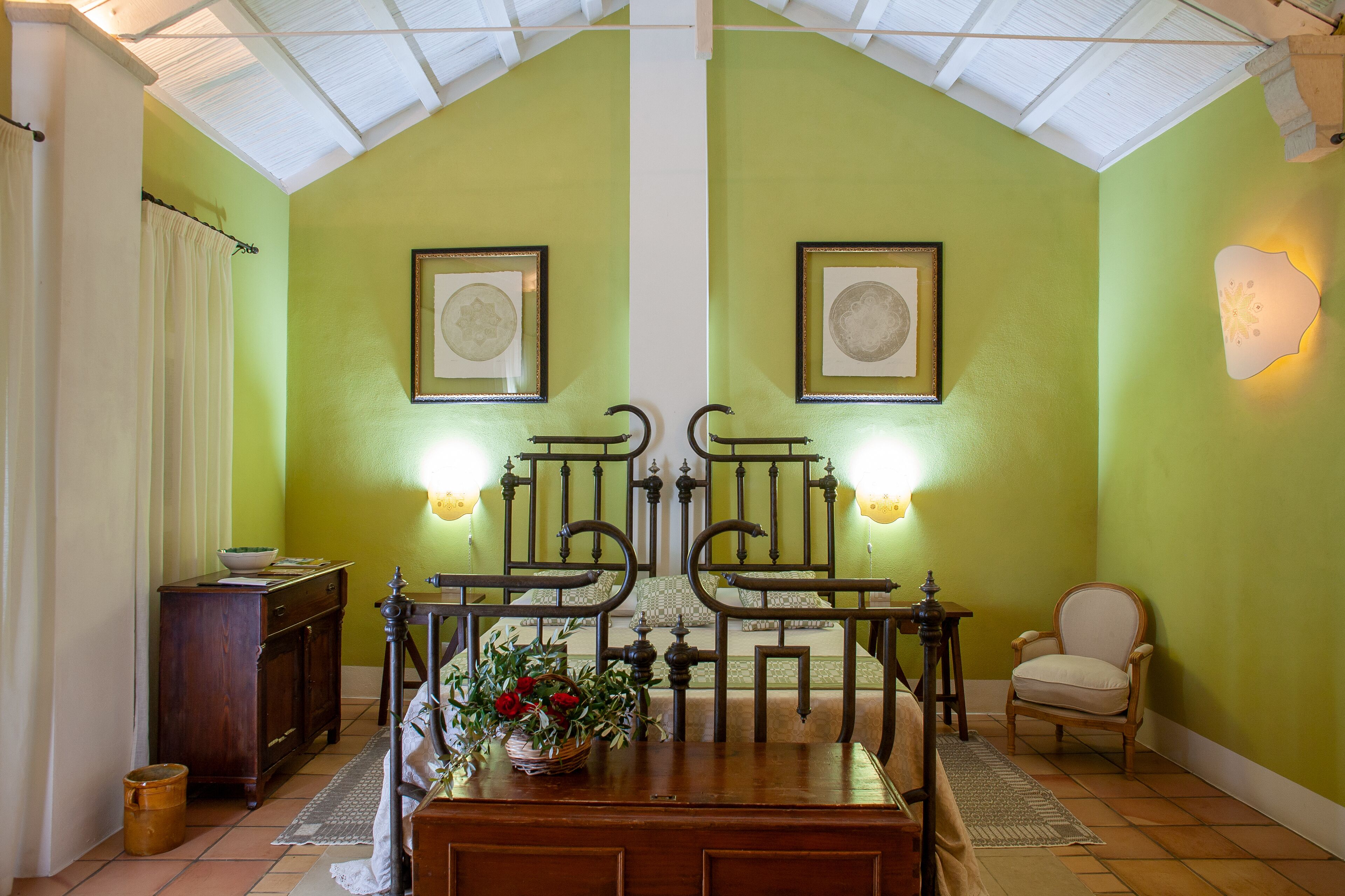 Una habitación con techo alto, paredes verdes, una cama con cabecera, dos sillas, una mesa con un florero y un pequeño armario.